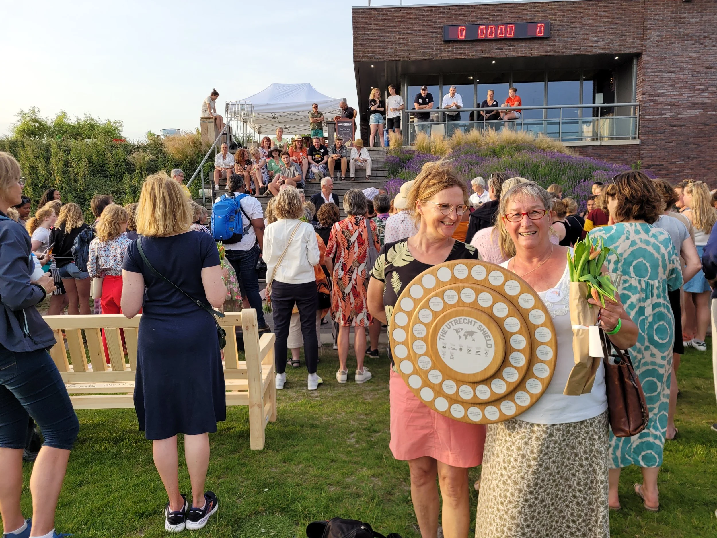 Two women smiling at a gathering outdoors, one holding a circular award and flowers, with a large crowd in the background.