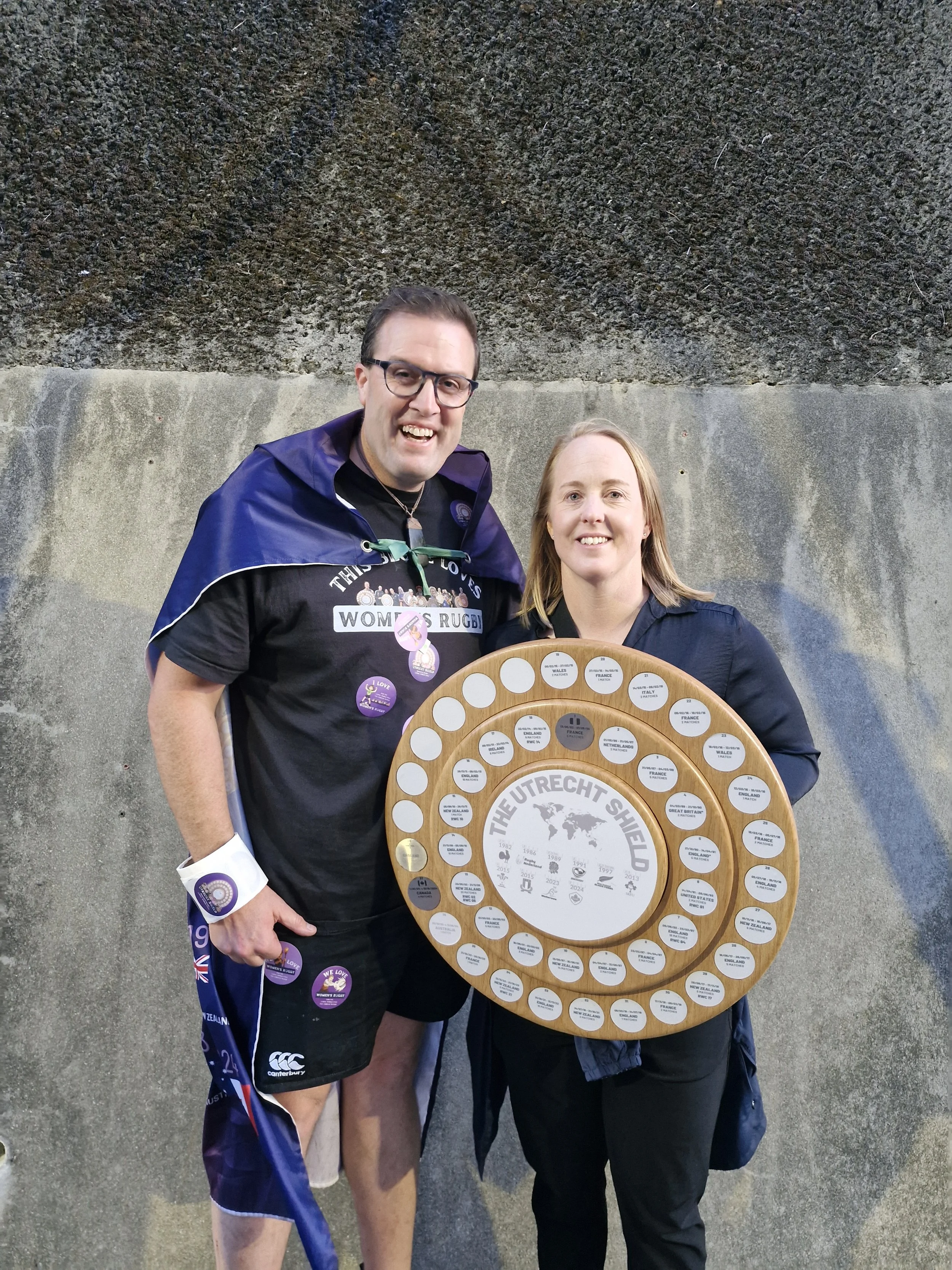 Two women standing next to each other, holding a large wooden circular plaque with smaller plaques around the edge, celebrating the British victory in rugby, with one woman dressed in rugby attire.