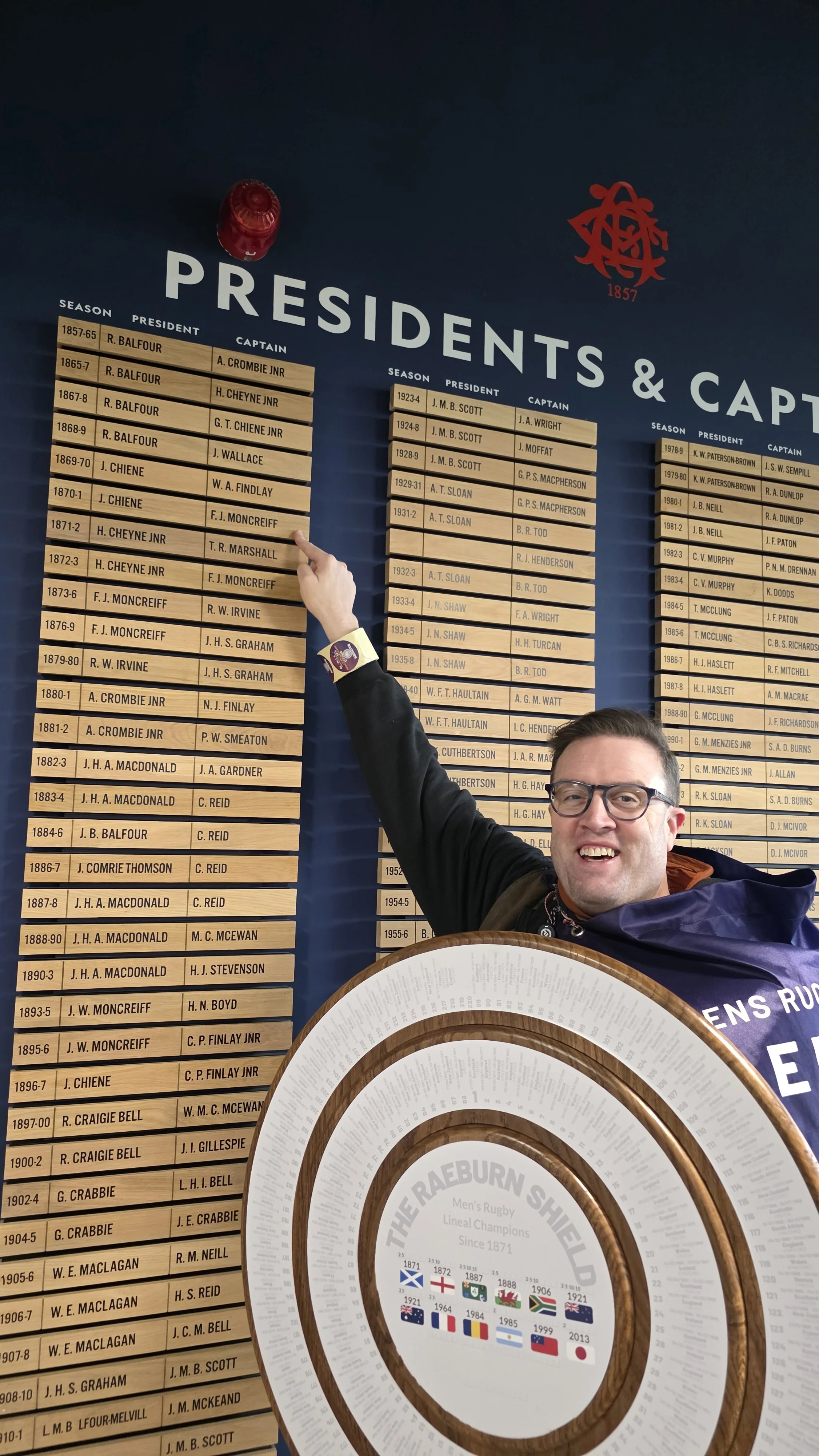 A man smiling and pointing at a wall display of rugby team captains and presidents, holding a large circular award or trophy in front of him.