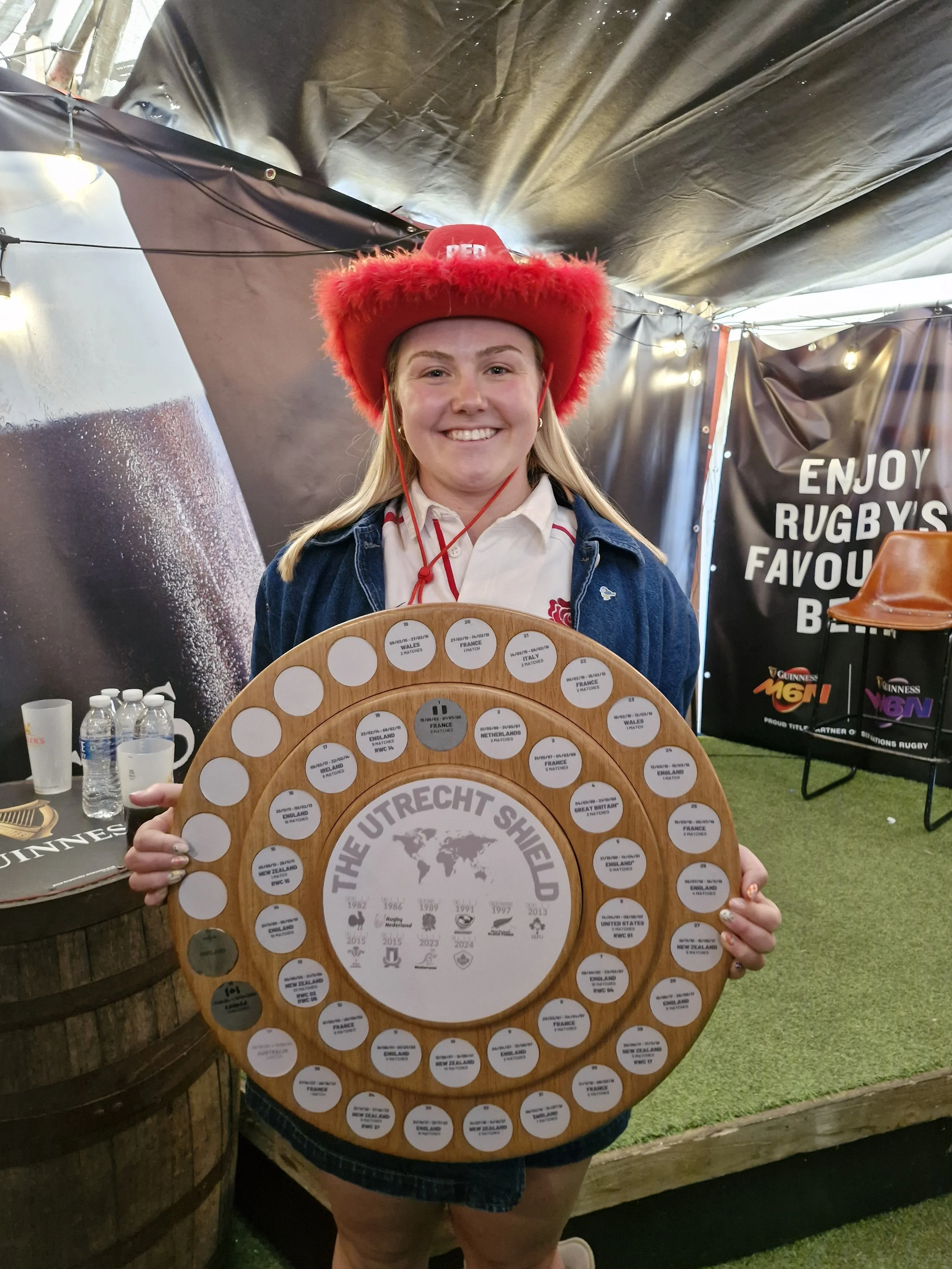 A young woman wearing a red furry hat and a denim jacket holding a large wooden rugby trophy, smiling for the camera. The background has a black curtain with text and bottles on a table.