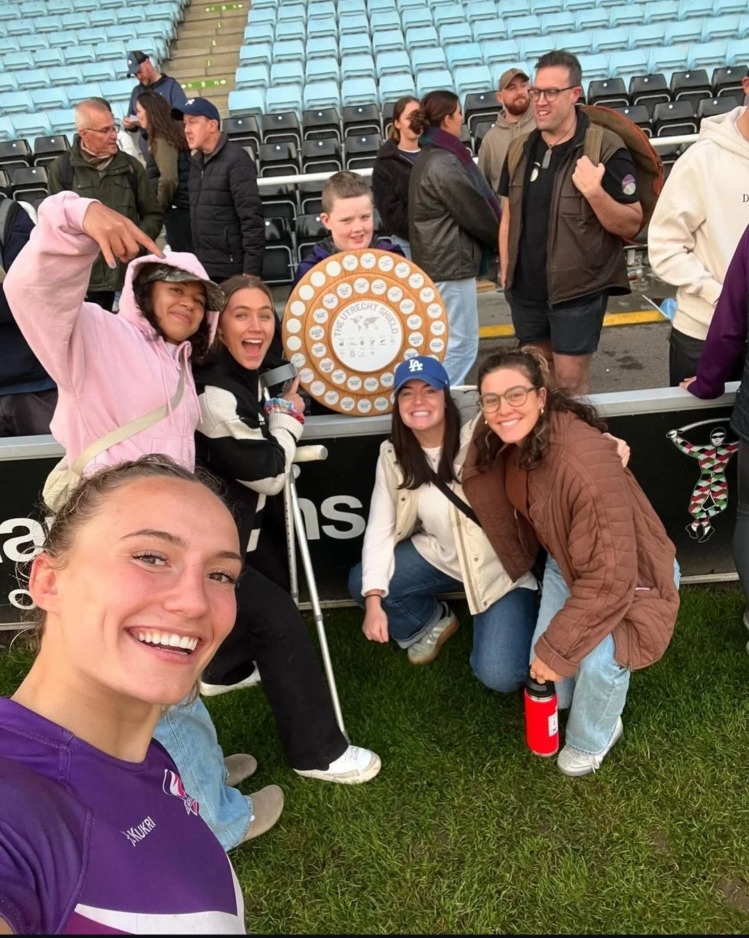 Group of smiling women and children posing for a selfie at a sporting event, with a trophy or award in the background and stadium seats behind them.
