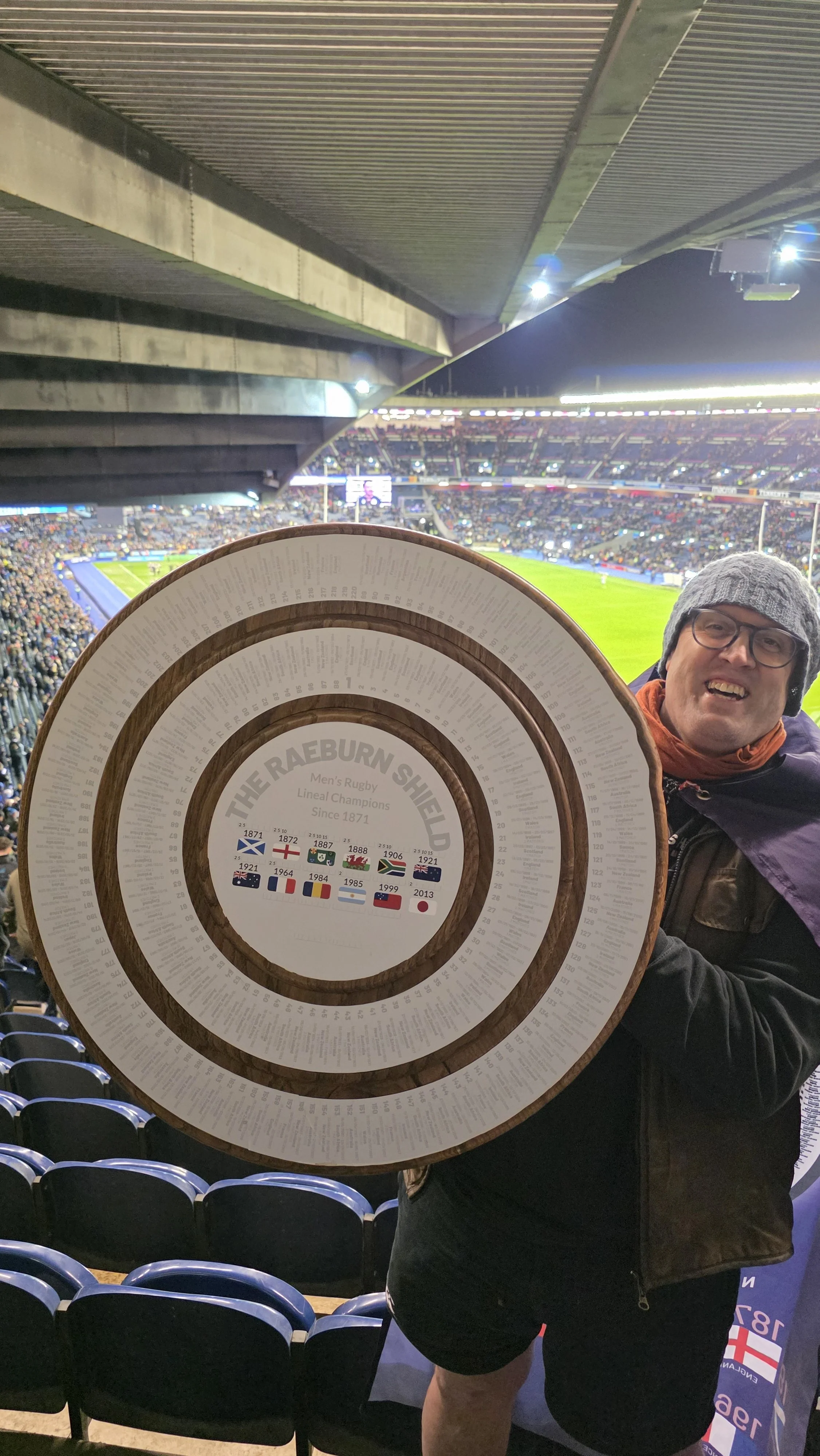 A man in glasses and a gray knit beanie smiling at the camera, holding a large circular commemorative plaque at a sports stadium with a rugby match in progress in the background.