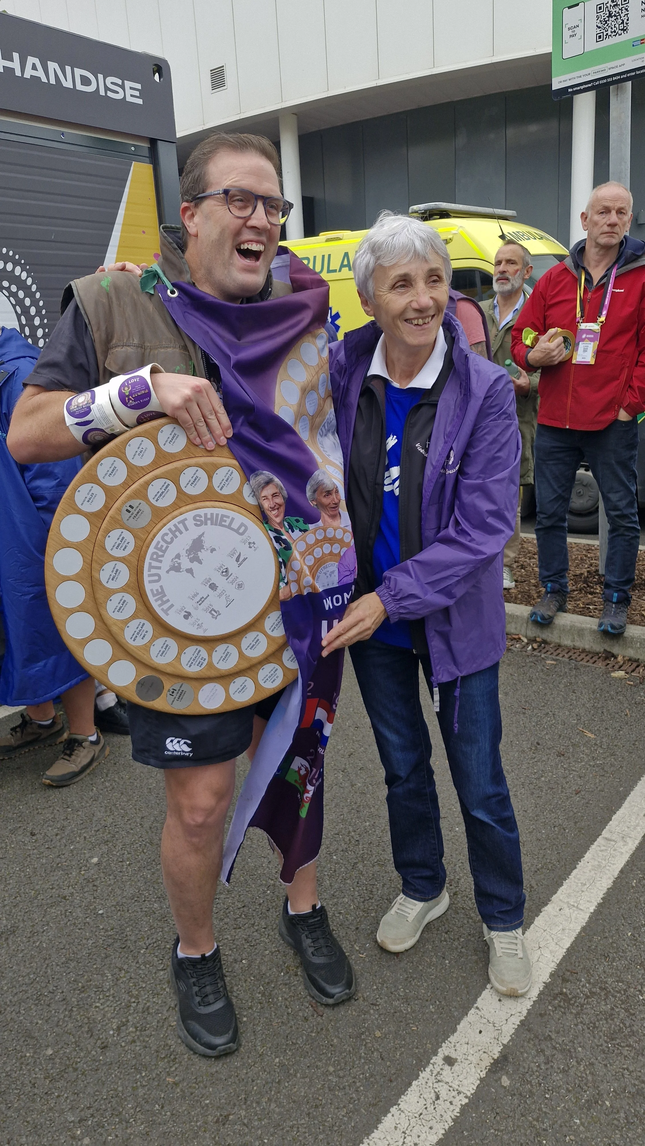 A man and a woman smiling and posing for a photo; the man is holding an award shaped like a wooden shield with multiple engraved plaques. They are outdoors, with people and emergency vehicles in the background.