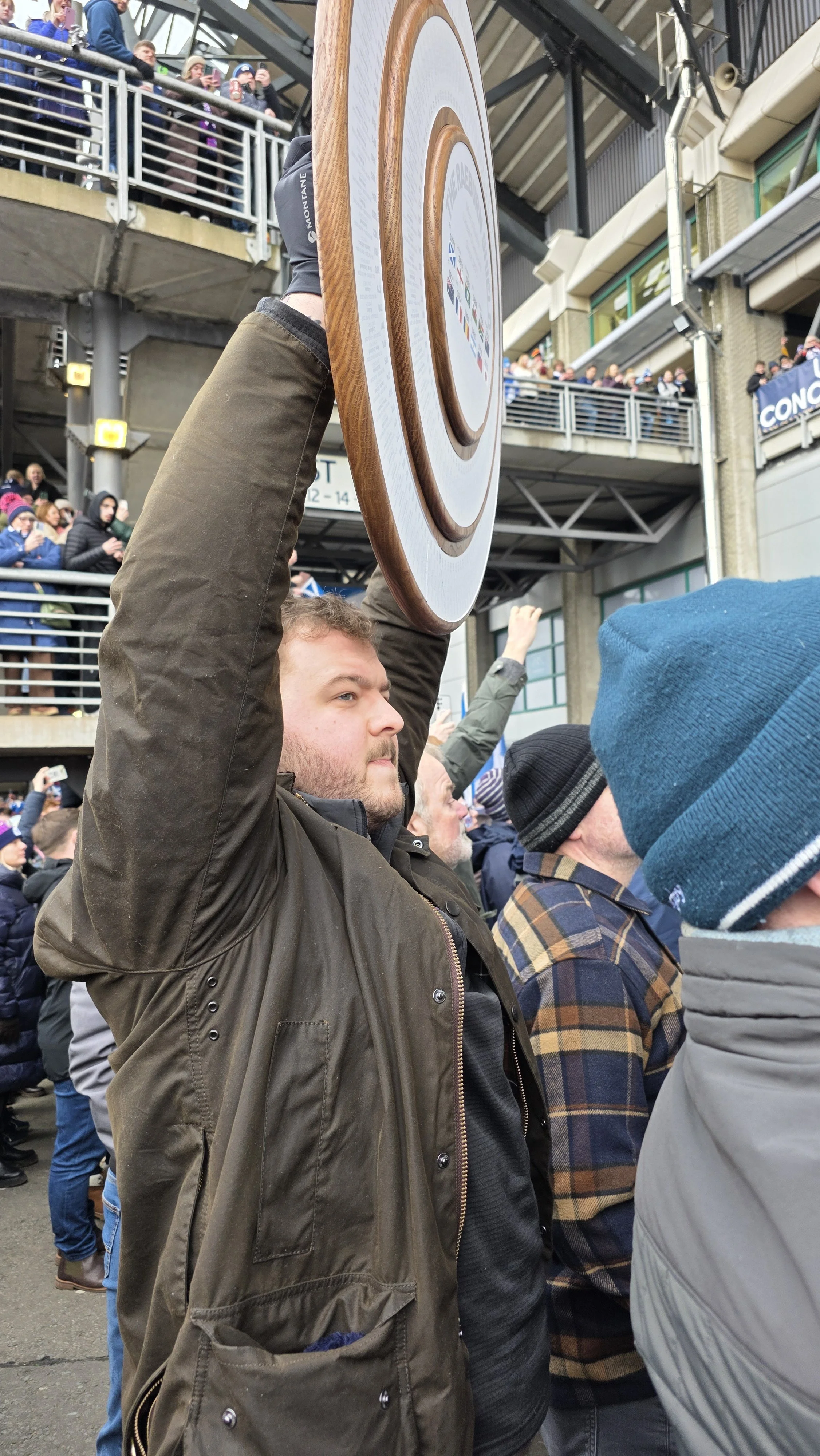 A man holding a large wooden sign at a crowded outdoor event during daytime.