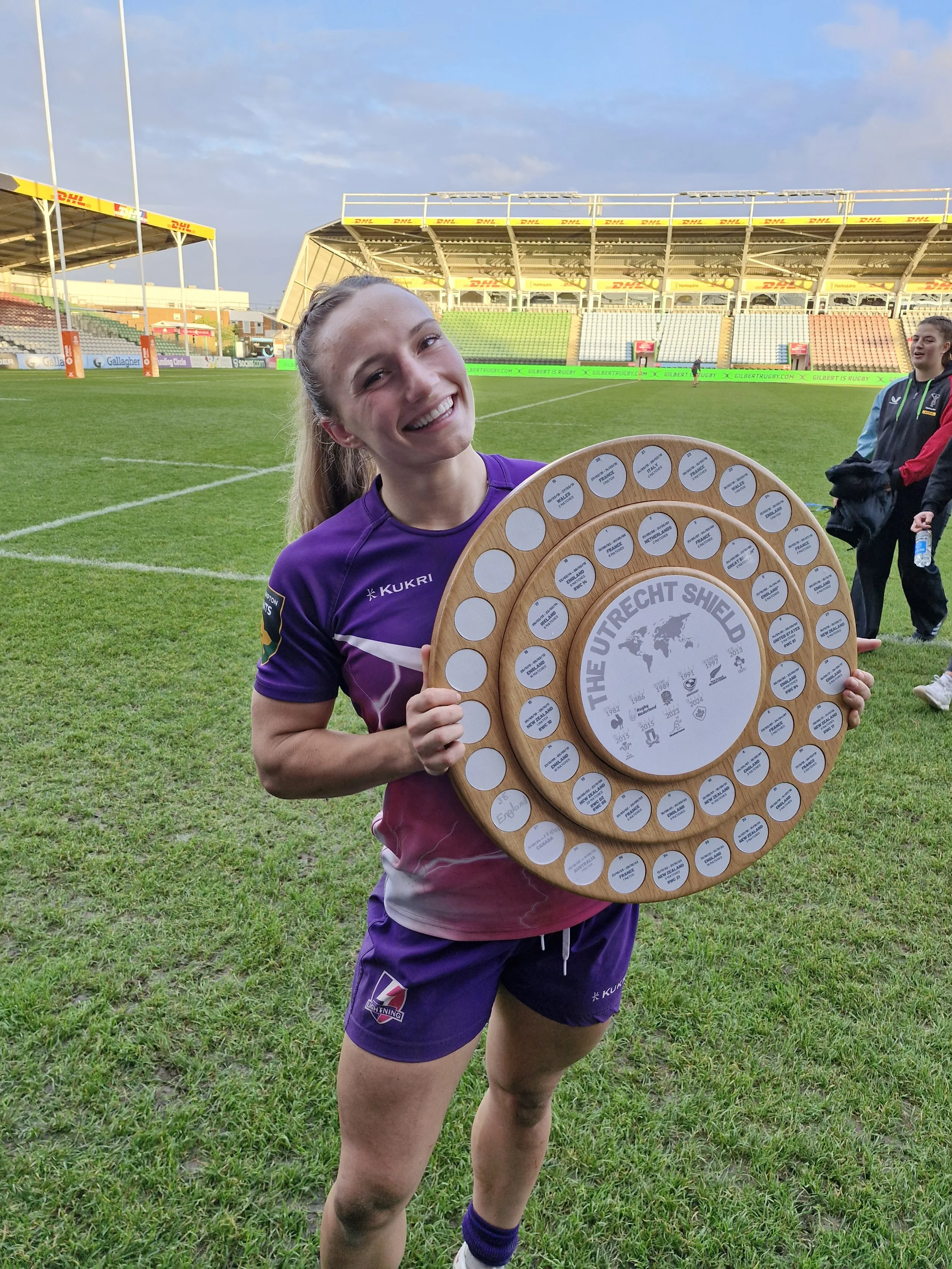 A female rugby player in purple uniform smiling and holding a circular wooden award with text and signatures, on a rugby field with empty stands in the background.