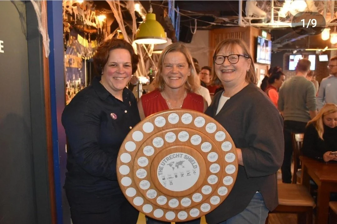 Three women smiling and holding a circular wooden plaque with multiple white labels, in a festive indoor setting with spider web decorations.