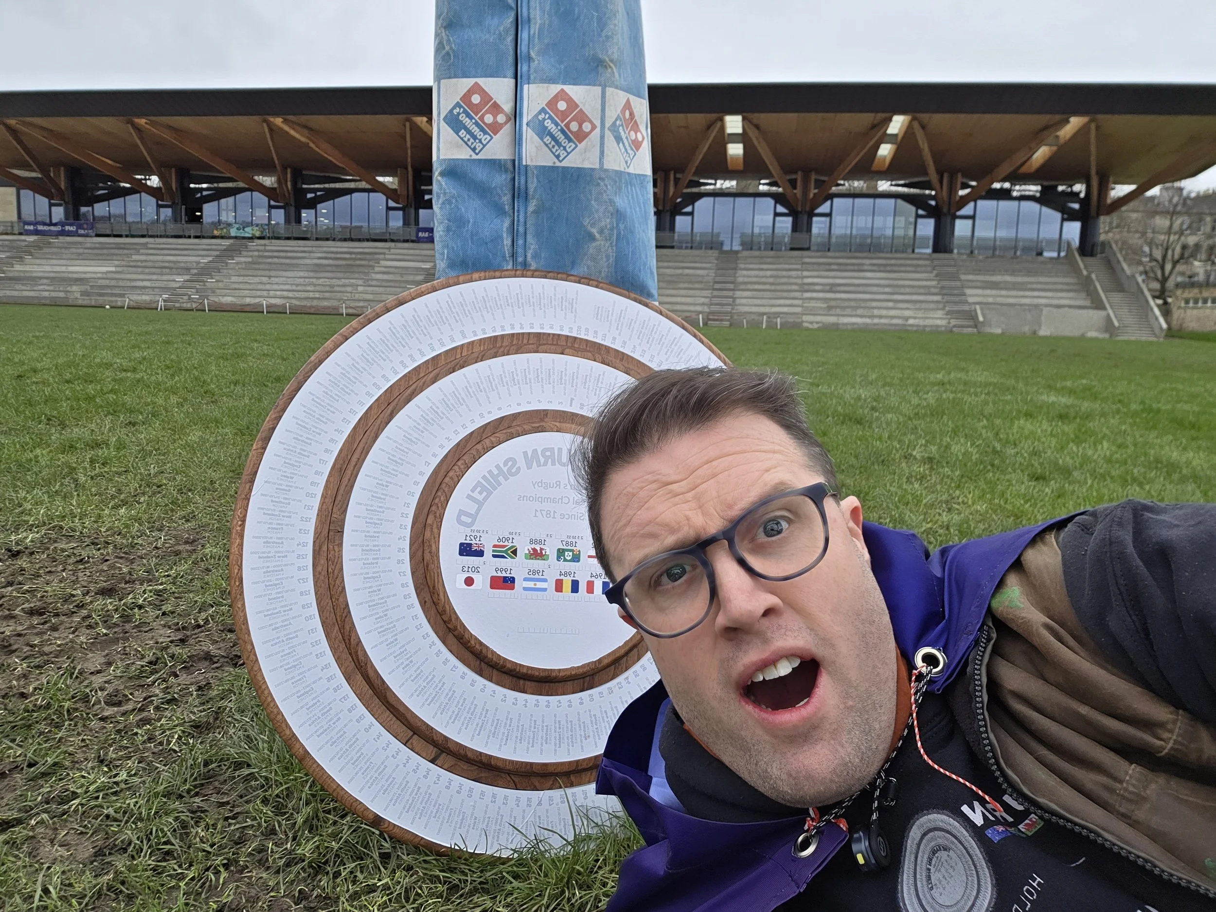 A man with glasses taking a selfie on the grass near a circular timeline display and a pole covered with Domino's pizza delivery bags at an outdoor stadium or arena.