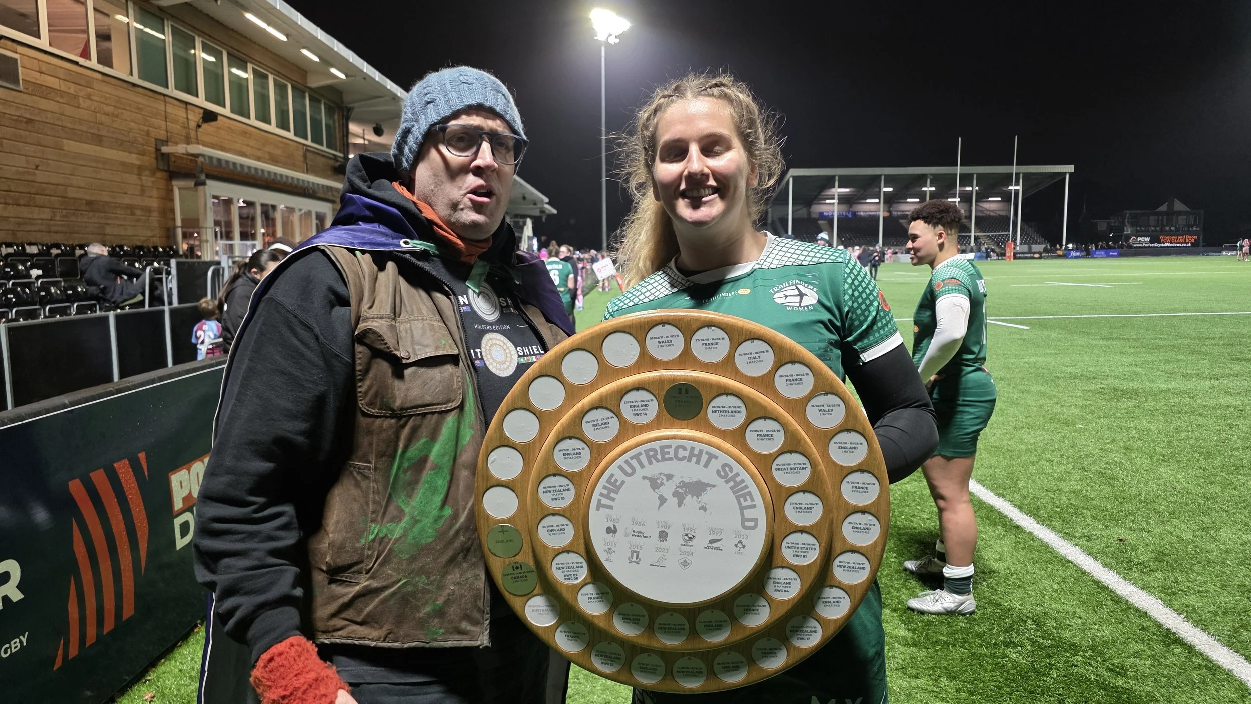 A woman in a green rugby uniform holding a wooden shield-shaped trophy at a rugby field, standing next to a man wearing glasses, a beanie, and a jacket, with other players in green uniforms in the background.