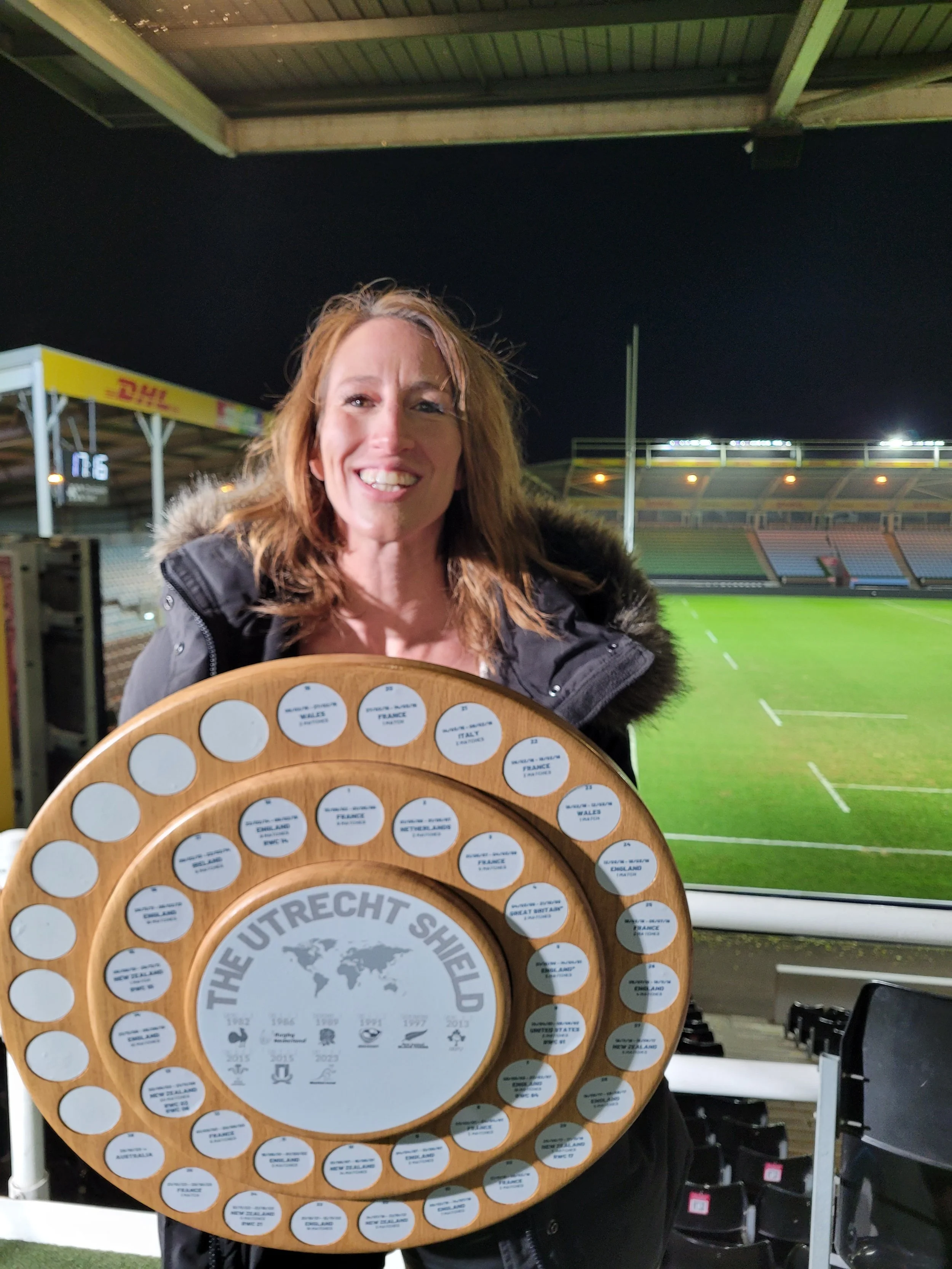 Woman smiling and holding a large wooden plaque with multiple circular labels, standing in a stadium at night.