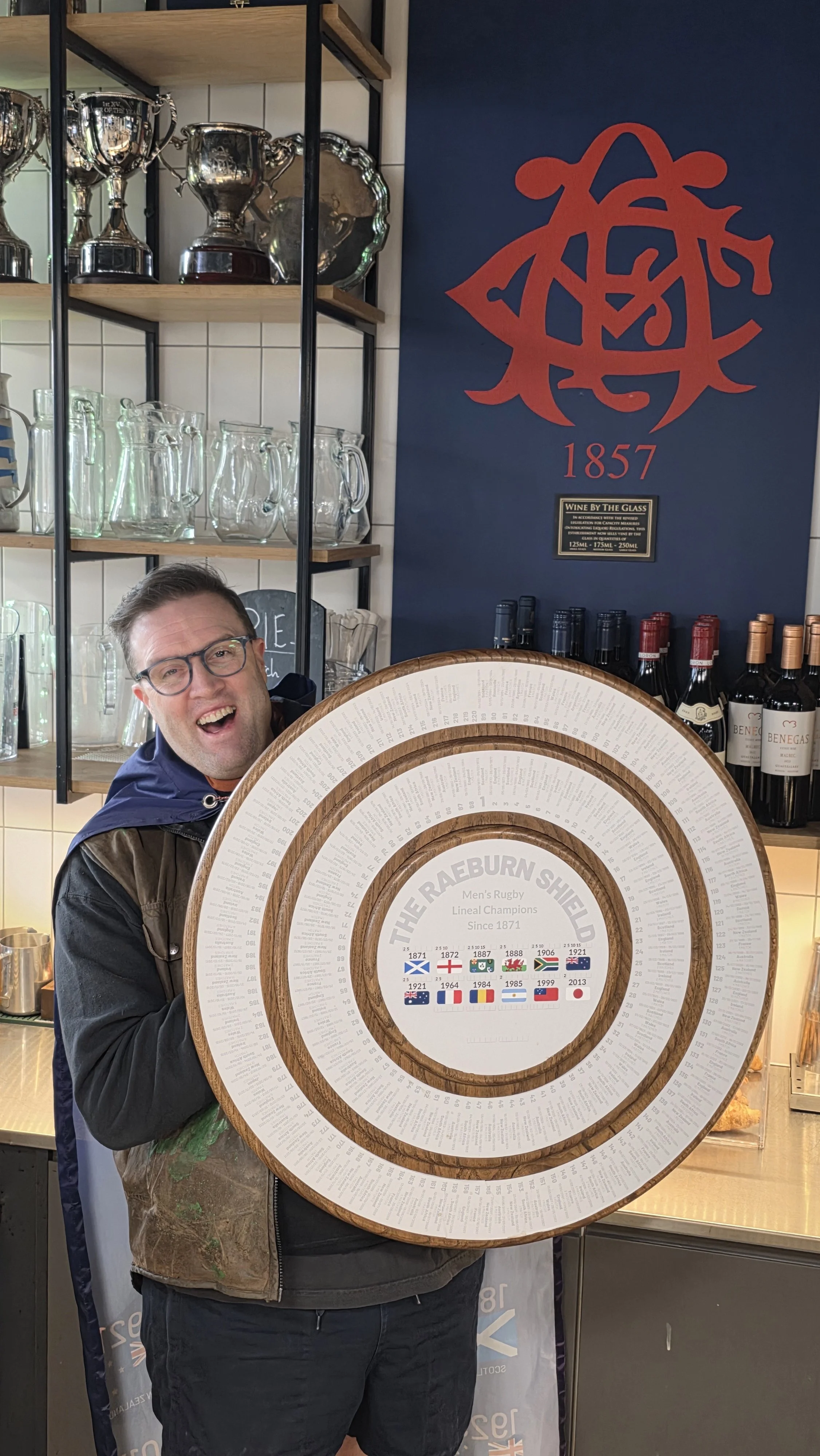 A man with glasses smiling and holding a large circular plaque with the Rugby Shield and flags of winning countries from 1871 to 2013, inside a room with shelves of glassware and bottles of wine.