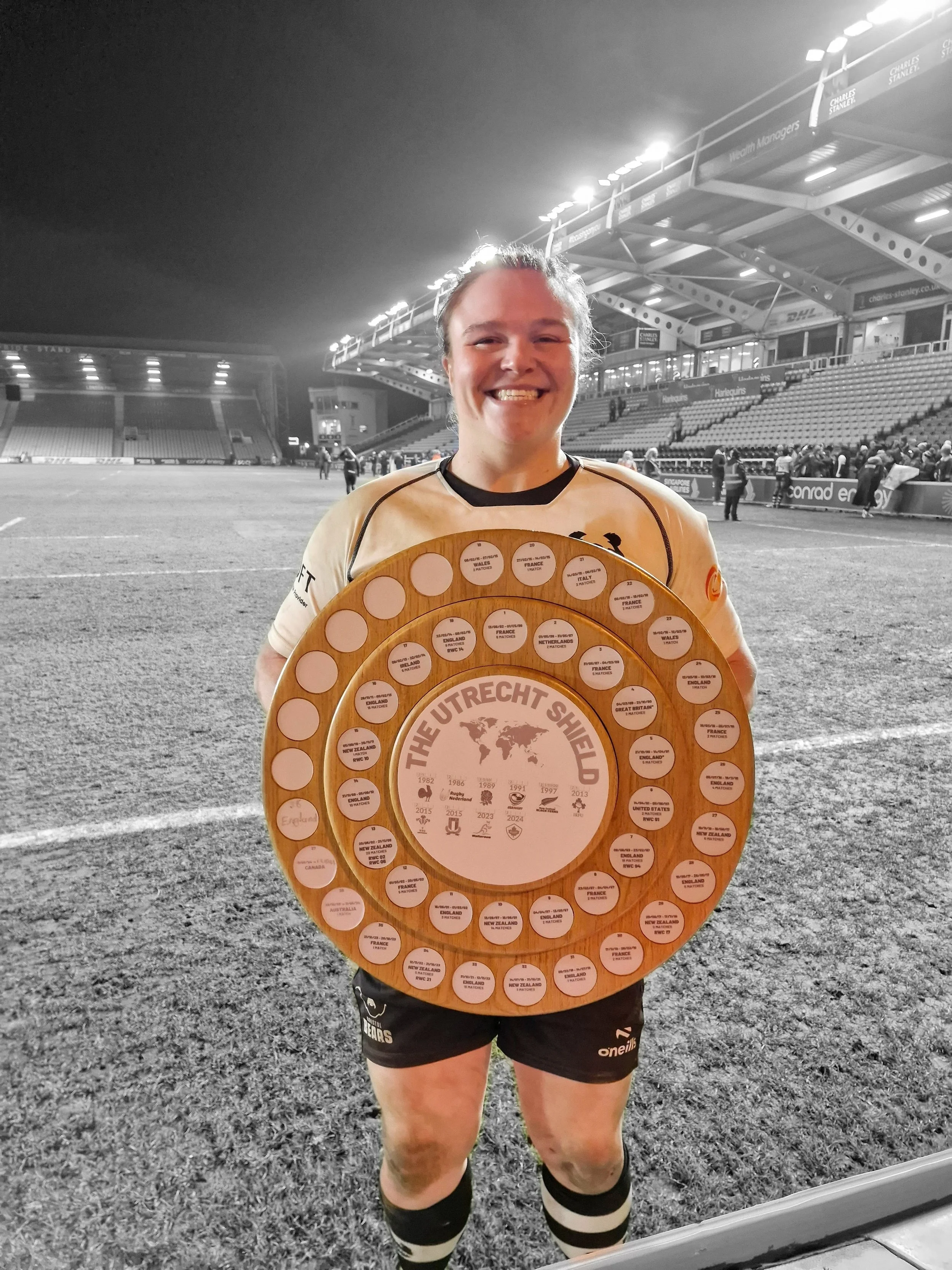 A woman rugby player smiling and holding a large wooden plaque with several small circular awards, standing on a rugby field at night with stadium stands and other people in the background.