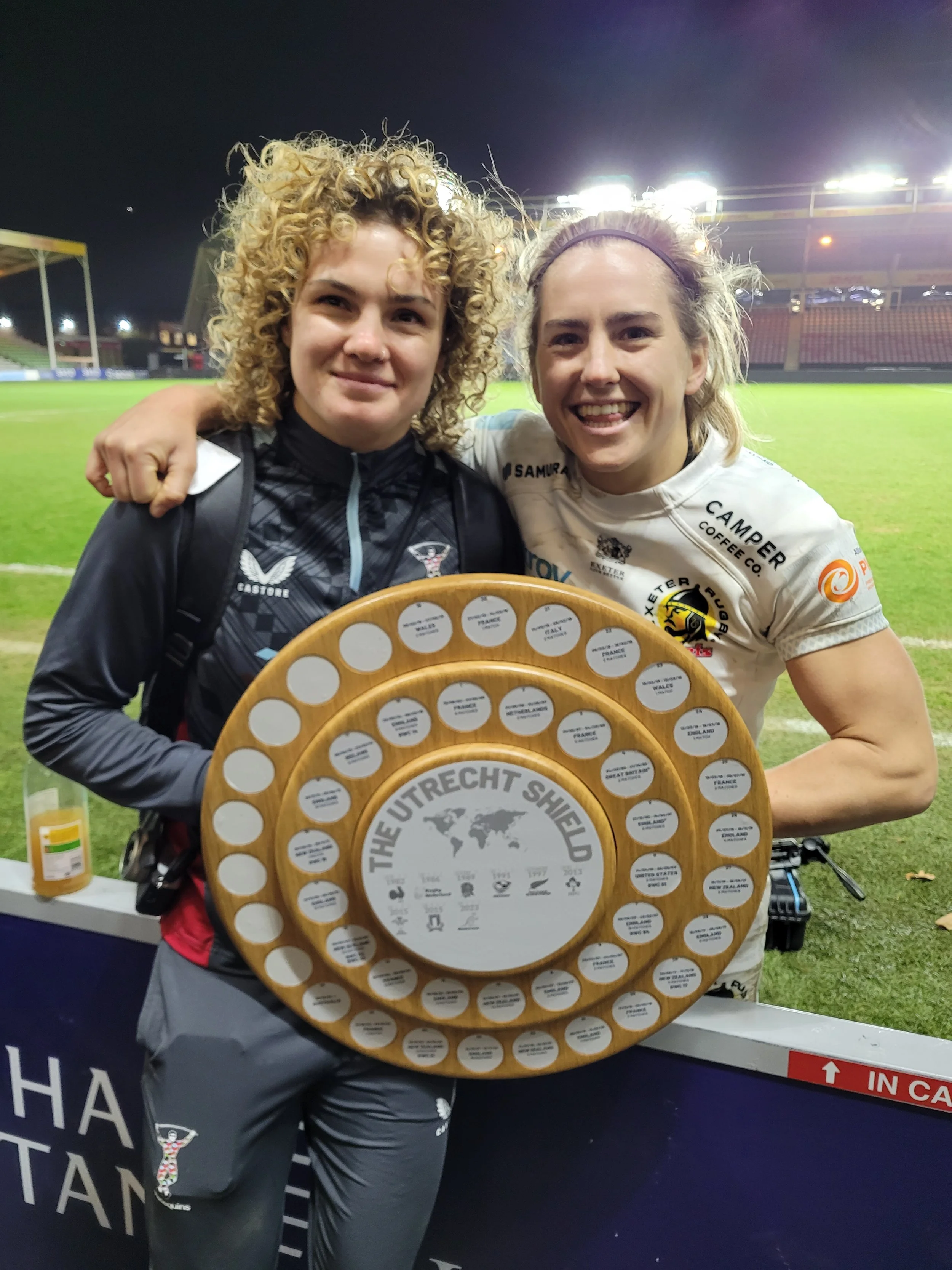 Two women holding a large wooden trophy on a sports field at night. The woman on the left has curly blonde hair and is wearing a black sports jacket; the woman on the right has blonde hair tied back with a headband, wearing a white rugby jersey. They