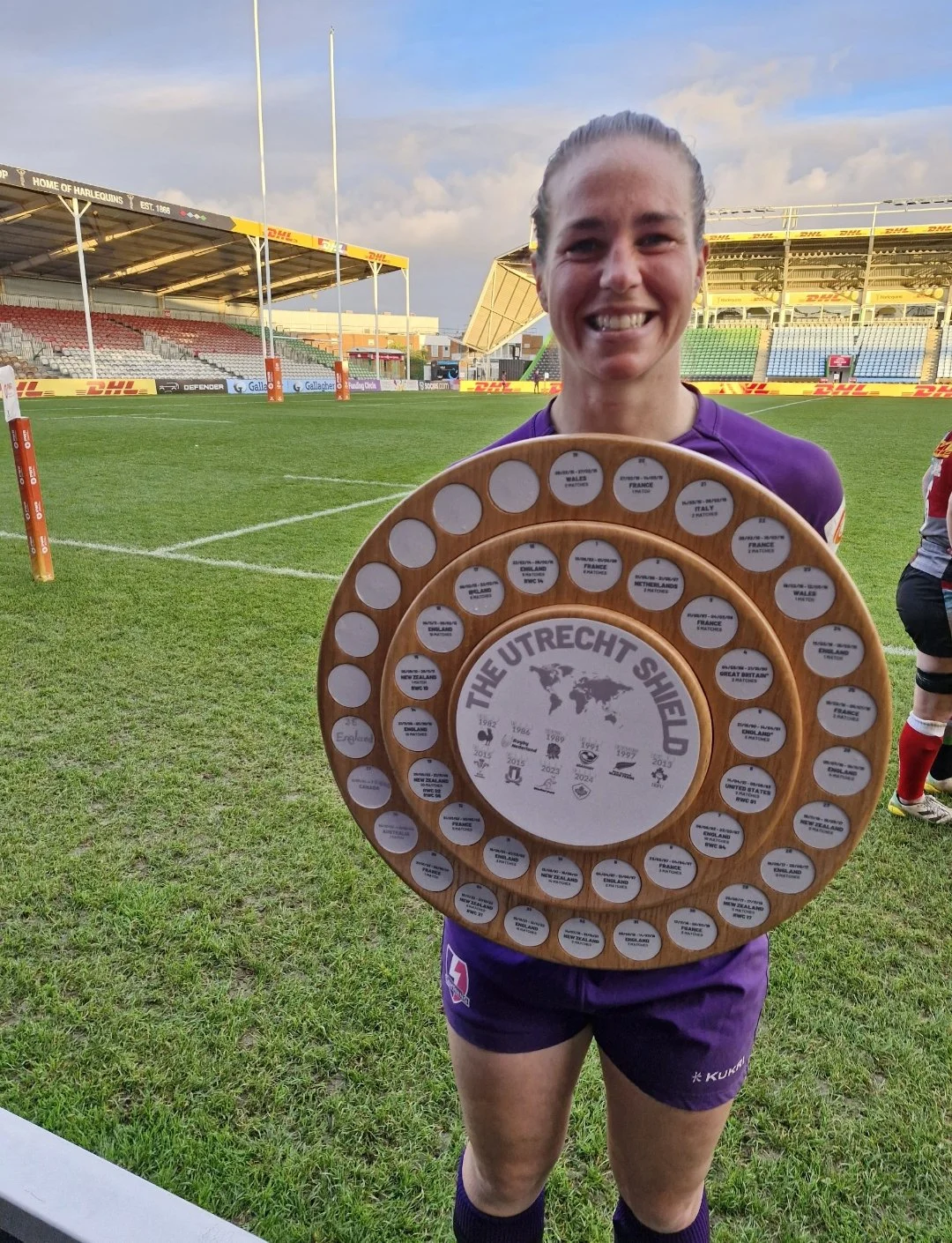 A female rugby player in a purple uniform smiling and holding a wooden plaque with multiple circular labels, standing on a rugby field with stadium seating and goalposts in the background.