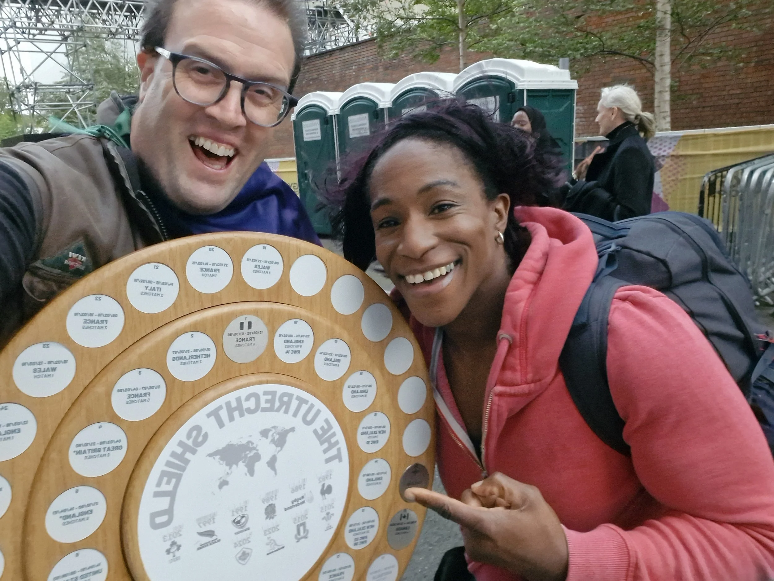 Two smiling people, a man and a woman, pointing at a circular wooden award labeled 'The Future Shield' and featuring a world map and logos, outdoors in front of portable toilets and a brick wall.
