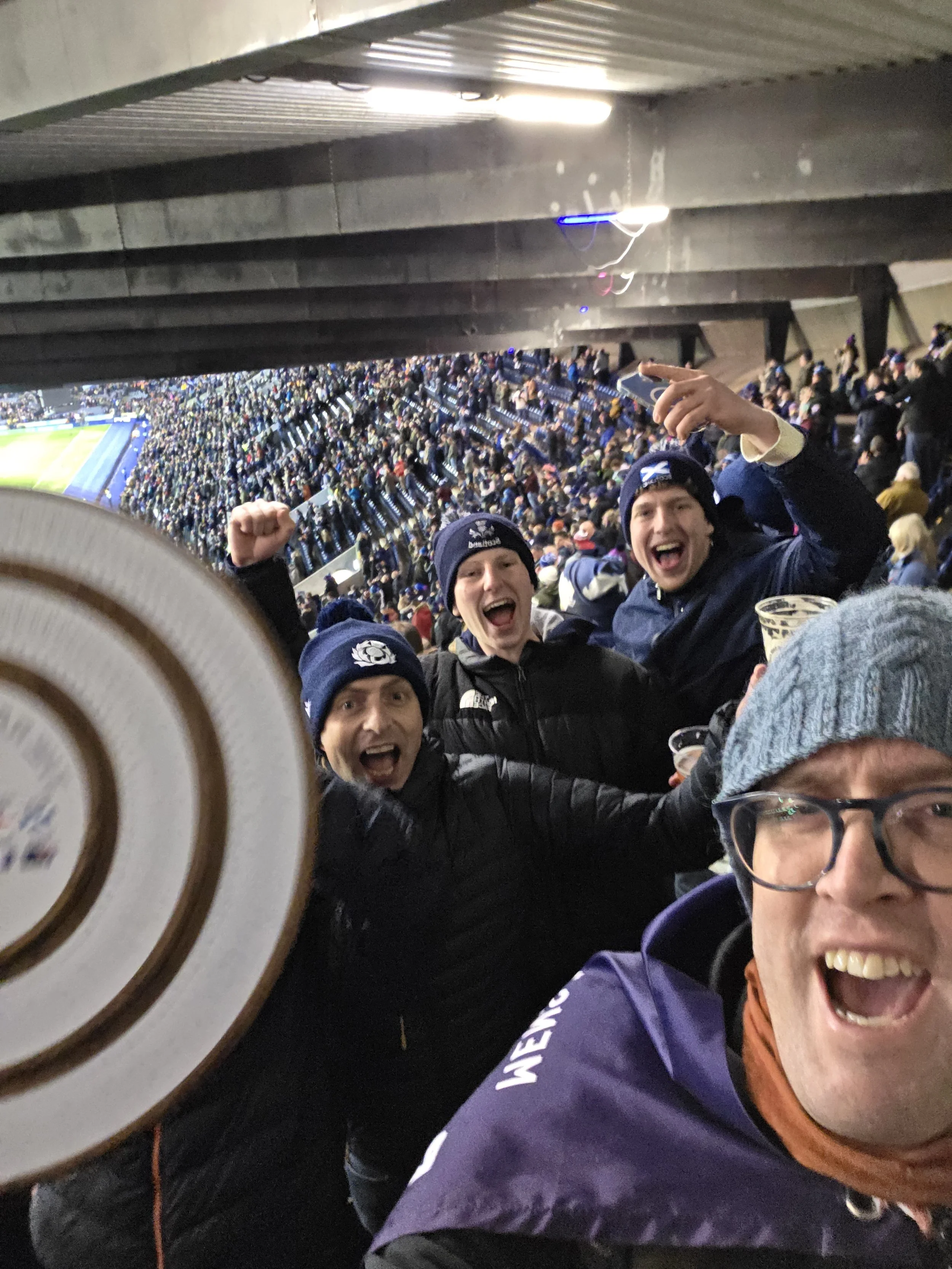 Group of excited soccer fans celebrating at a stadium, with a large crowd in the background, some holding drinks, under a roofed section.