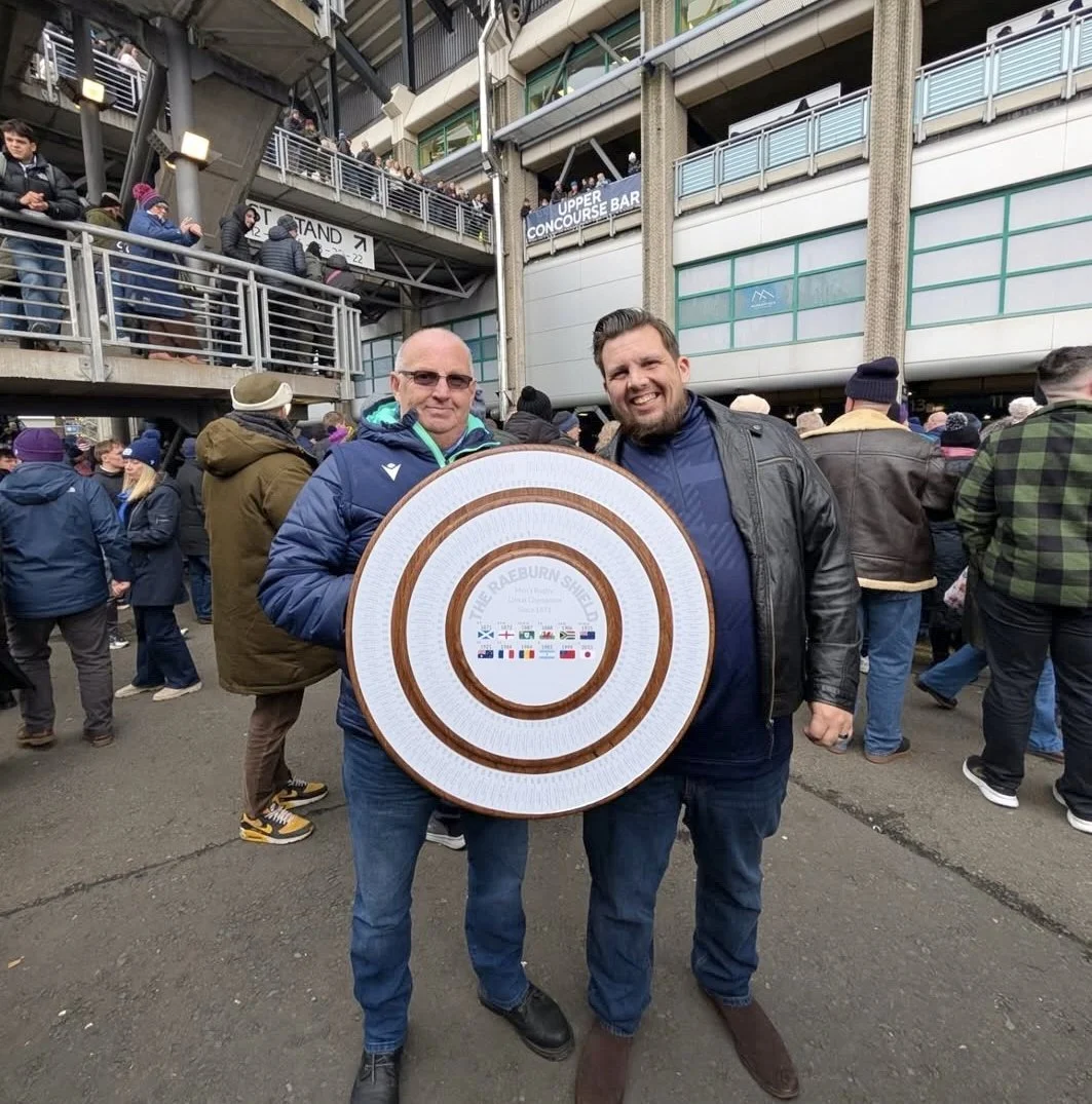 Two men posing outdoors surrounded by a crowd at a sports stadium, holding a circular wooden plaque with the words "The Raeburn Shield" and various flags printed on it.