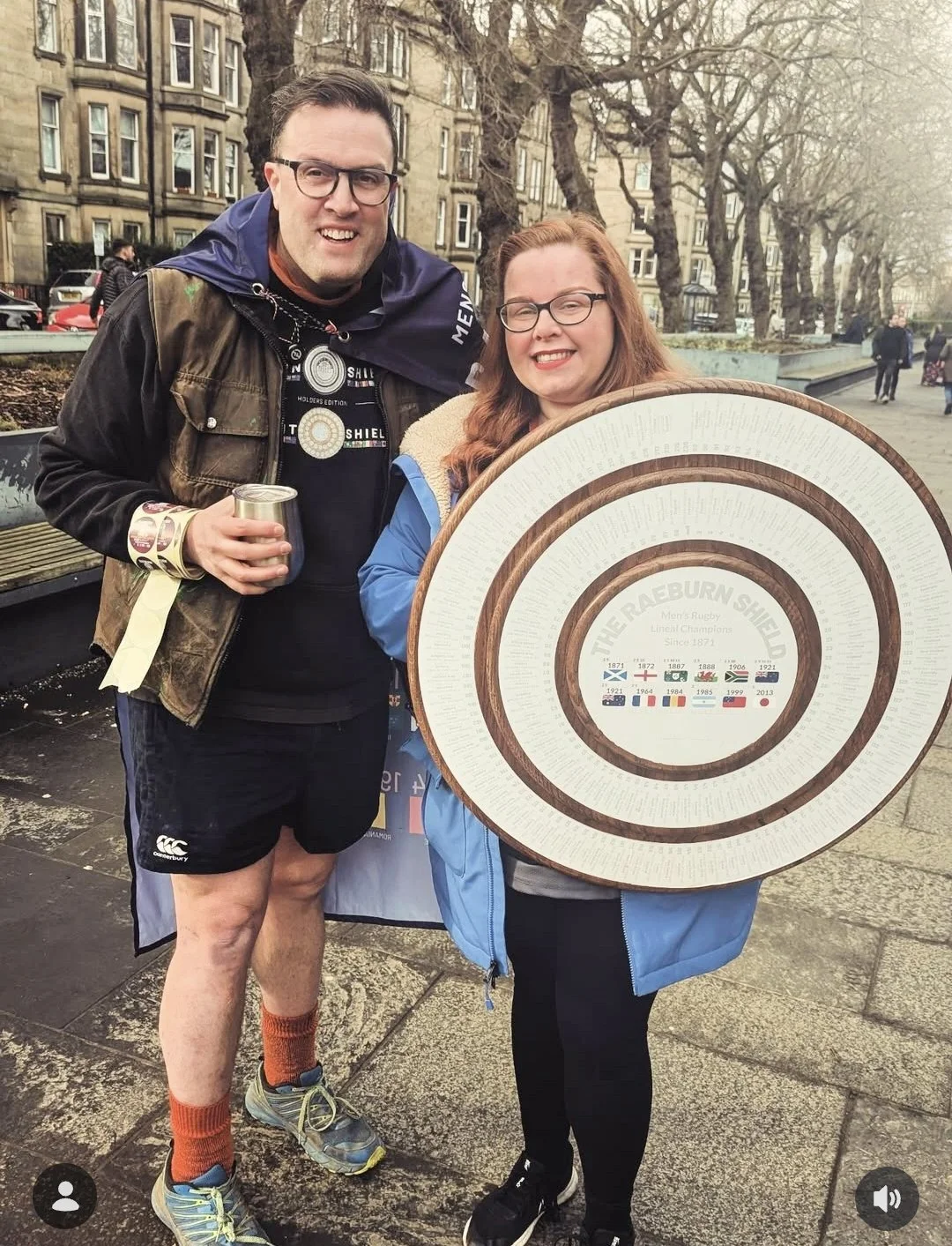 A man and woman standing outdoors in a city park, holding a large circular rugby shield with multiple concentric rings and historical dates and flags. The man is wearing a brown jacket, black shorts, orange socks, and glasses, holding a drink. The wo
