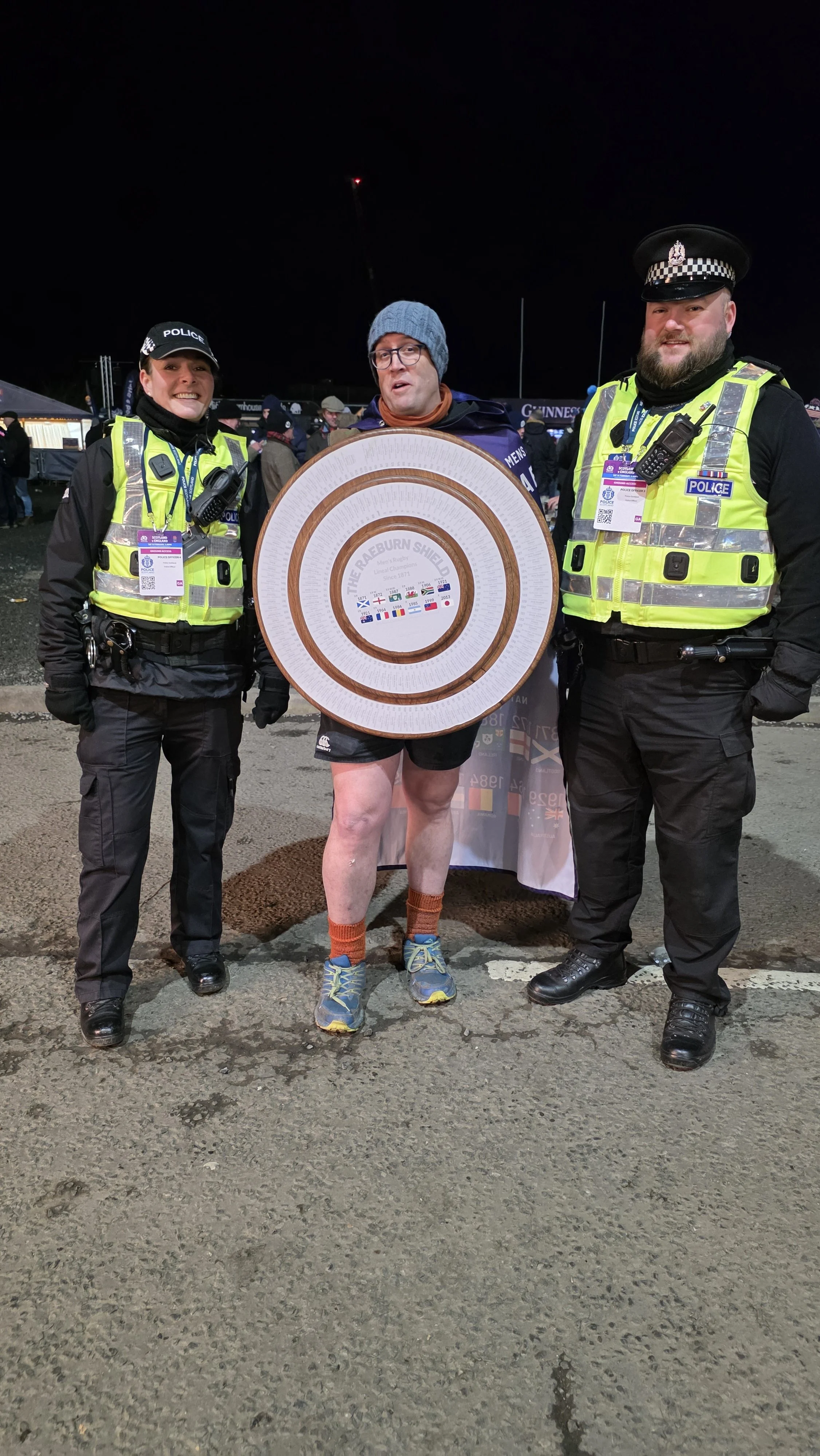 A man with a shield and two police officers standing outdoors at night. The man is wearing shorts, a beanie, glasses, and tennis shoes, and is holding a shield labeled 'The Raeburn Shield.' The police officers are in uniform and high-visibility vests