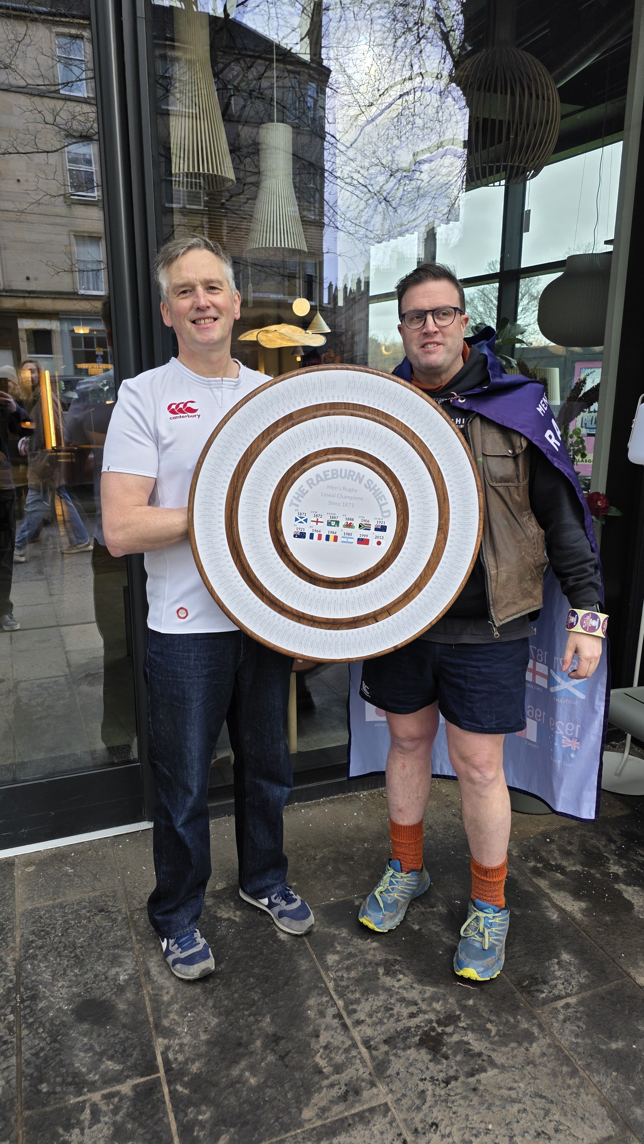 Two men standing outside in front of a glass storefront, holding a large circular framed piece with a map and text commemorating The Raeburn Shield, with reflections of buildings and trees in the glass.
