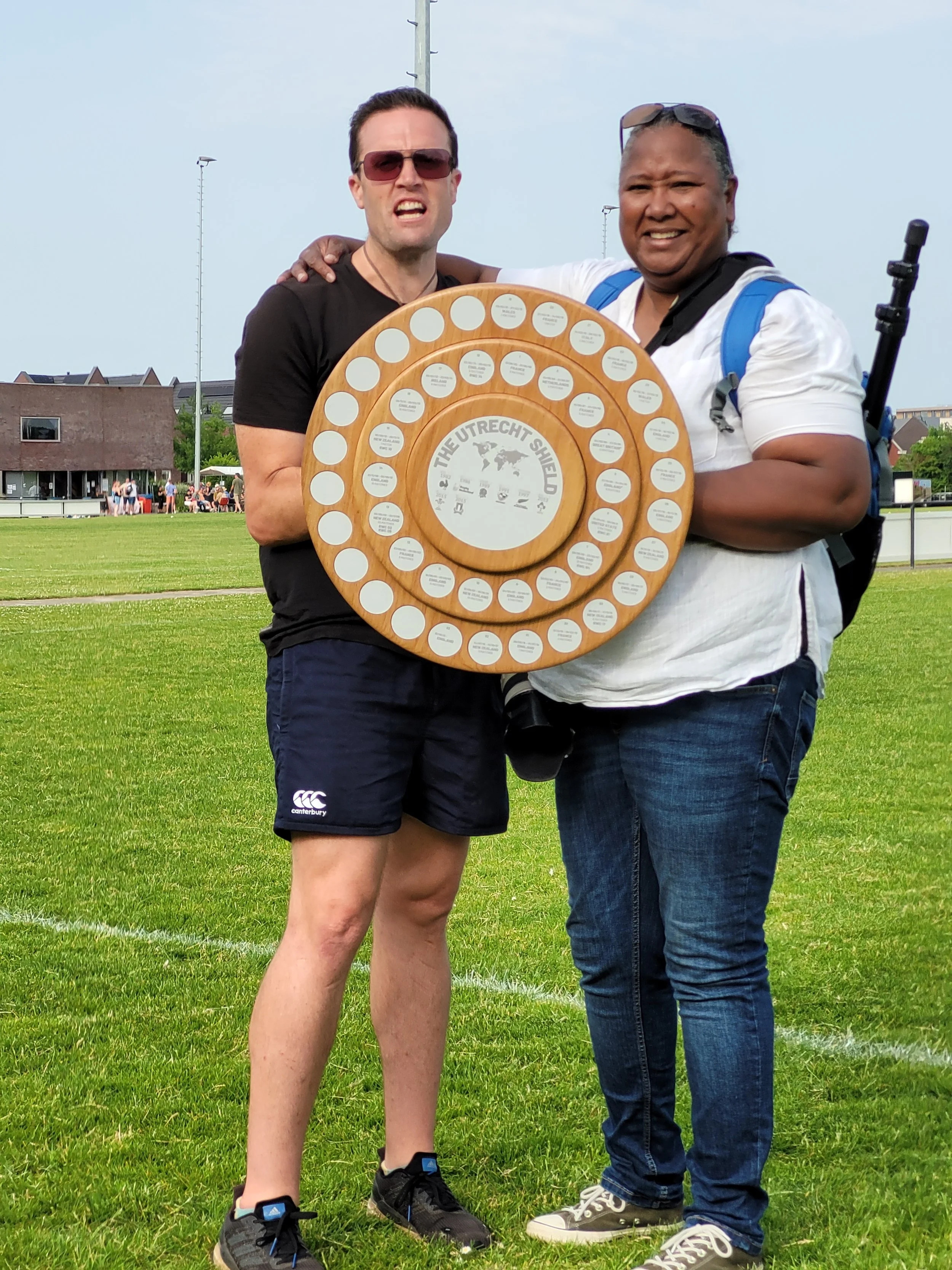 Two men standing on a grassy field holding a circular wooden trophy with white circular labels. One man is wearing a black t-shirt, shorts, and sunglasses, and the other is wearing a white shirt, jeans, and glasses on his head. They are smiling and t
