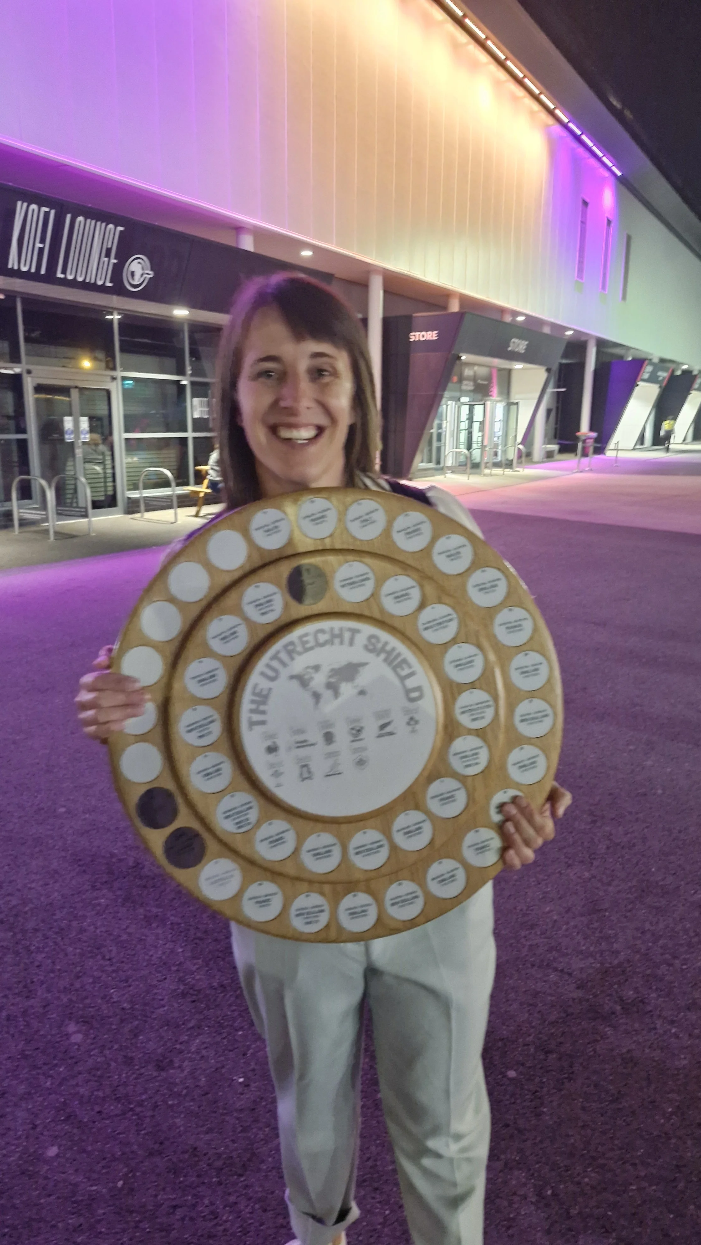 A woman holding a large circular wooden plaque with the words "The Utrecht Shield" and a map of the world engraved on it, outside a modern building with colorful LED lighting.