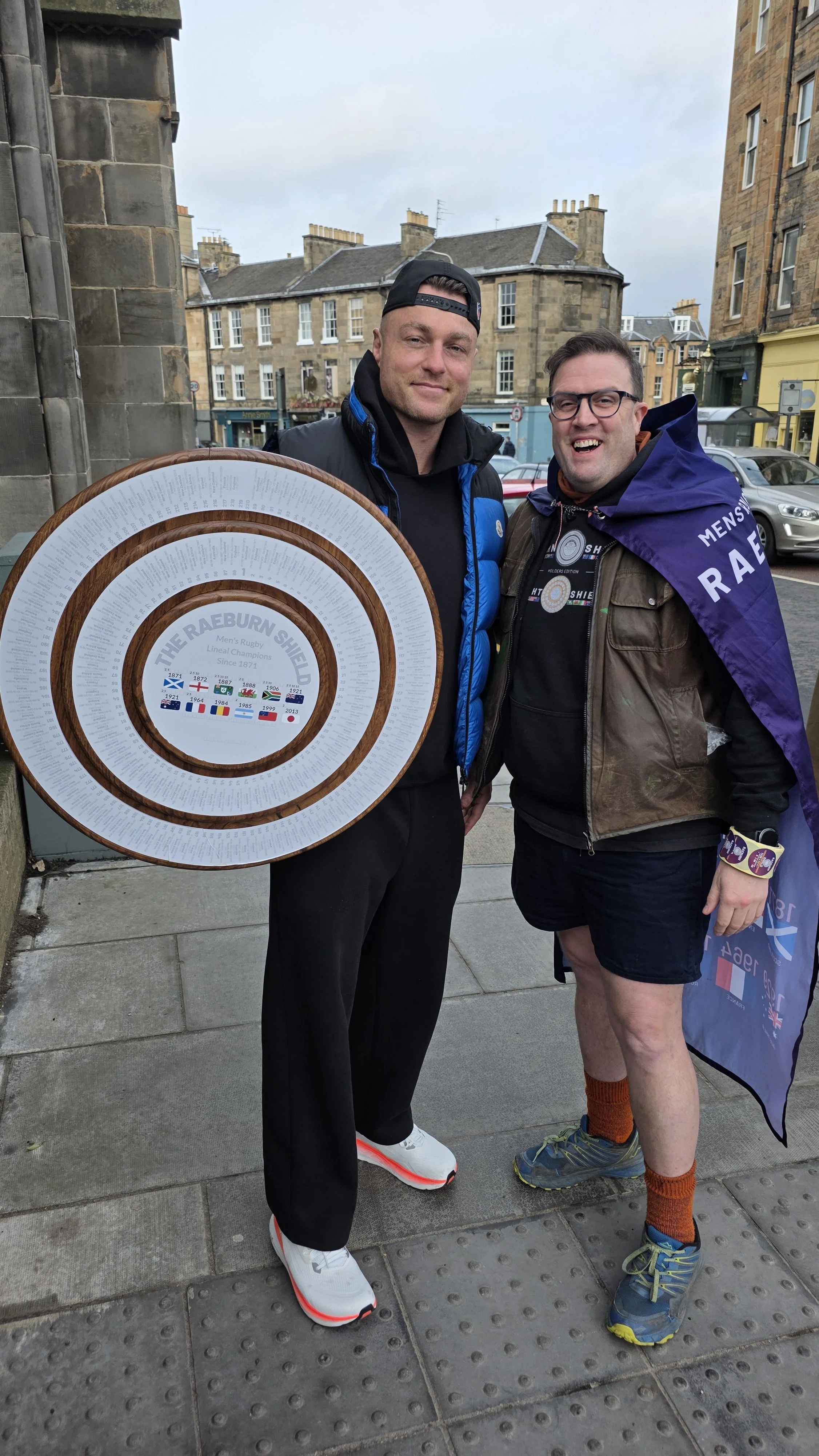 Two men standing outdoors, one holding a large round framed item, both smiling, with buildings and cars in the background.