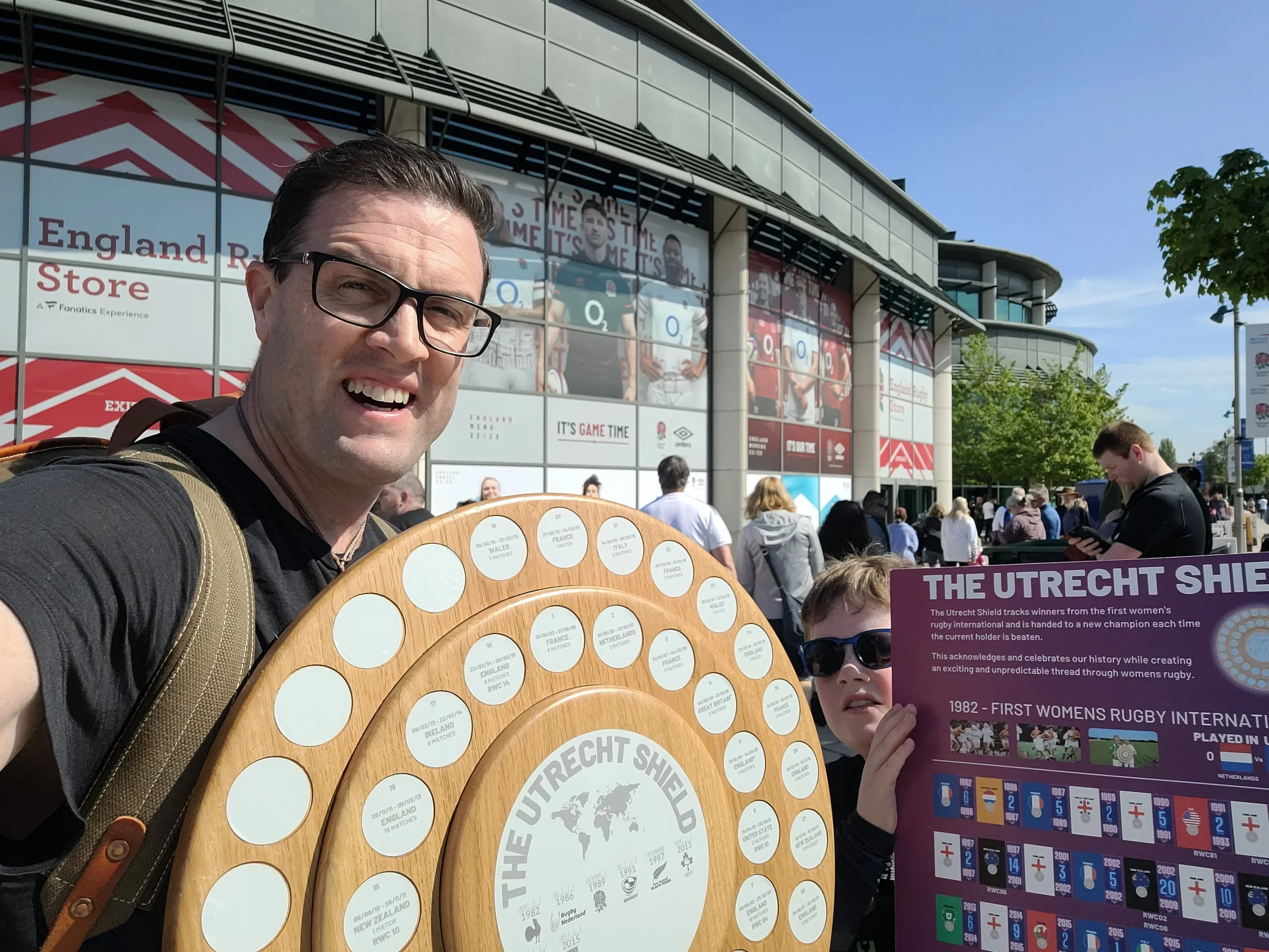 A man wearing glasses and a backpack taking a selfie outside a stadium with a woman holding an informational sign about the Utrecht Shield. The stadium has large banners showing rugby players and text celebrating women's rugby.