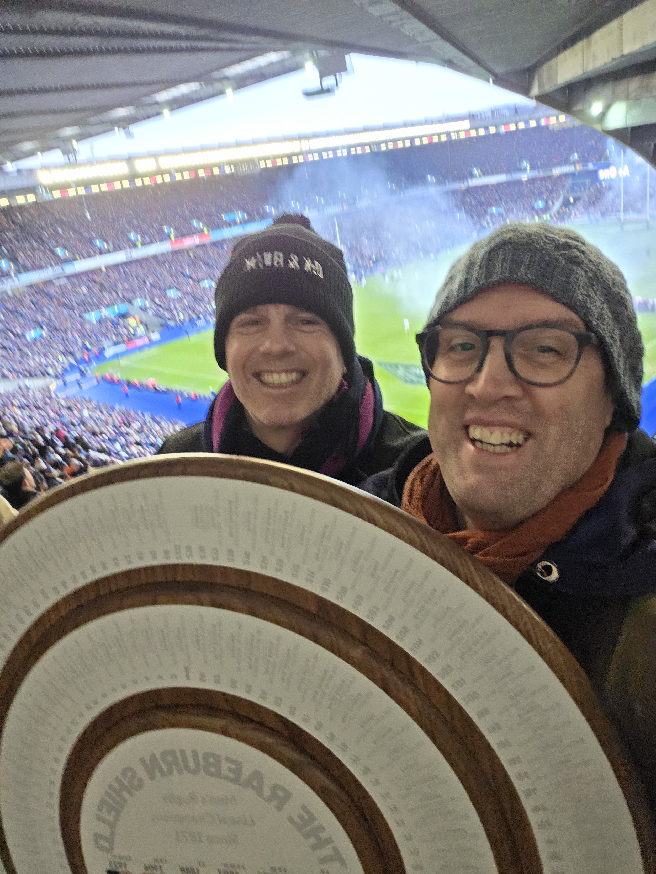 Two smiling men in winter clothing and hats at a stadium, holding a large circular menu or brochure, with a packed sports stadium and crowd in the background.
