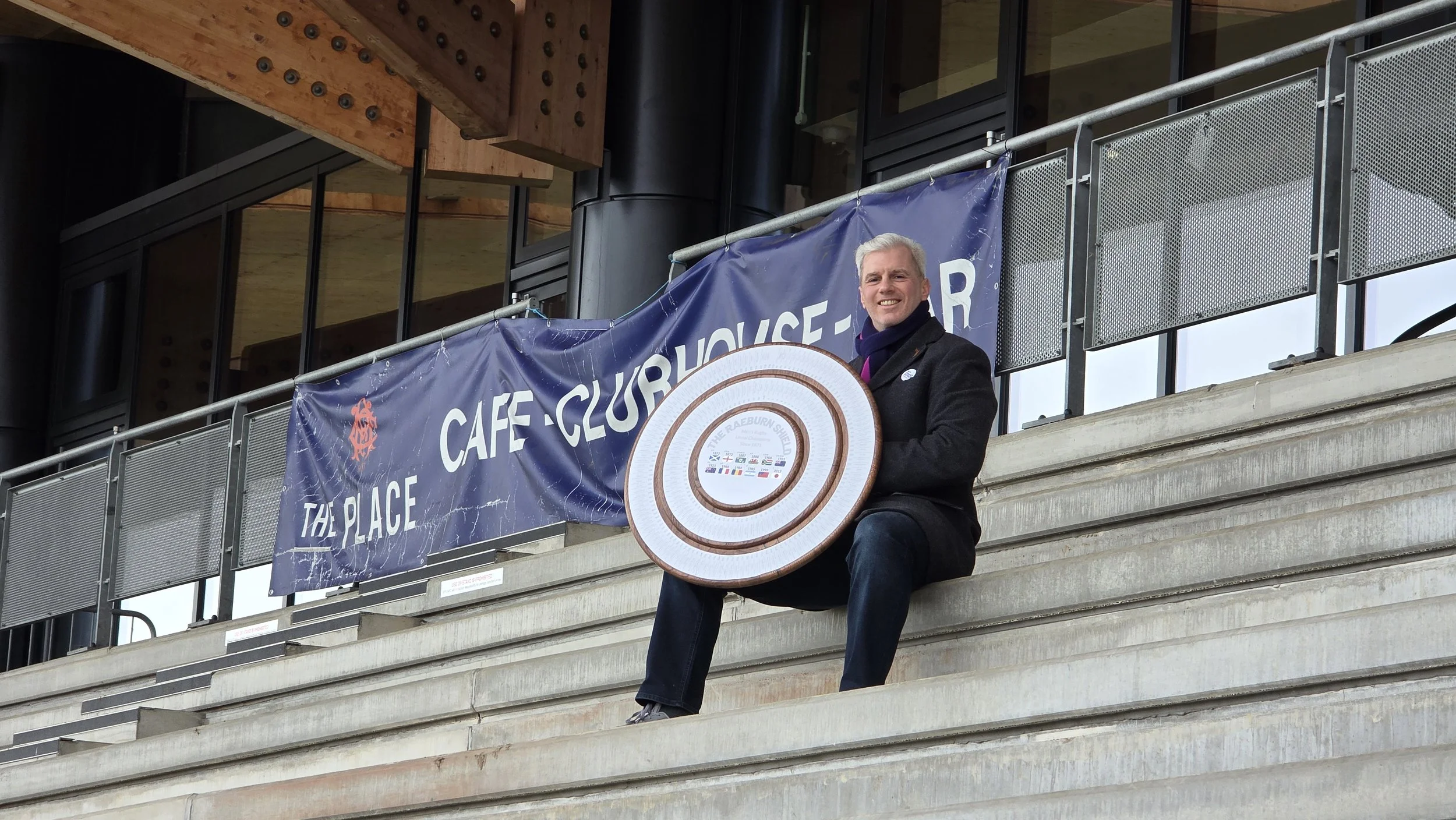 A man sitting on the stairs outside a building holds a circular plaque or award. Behind him is a banner that says 'The Place Cafe - Clubhouse.'