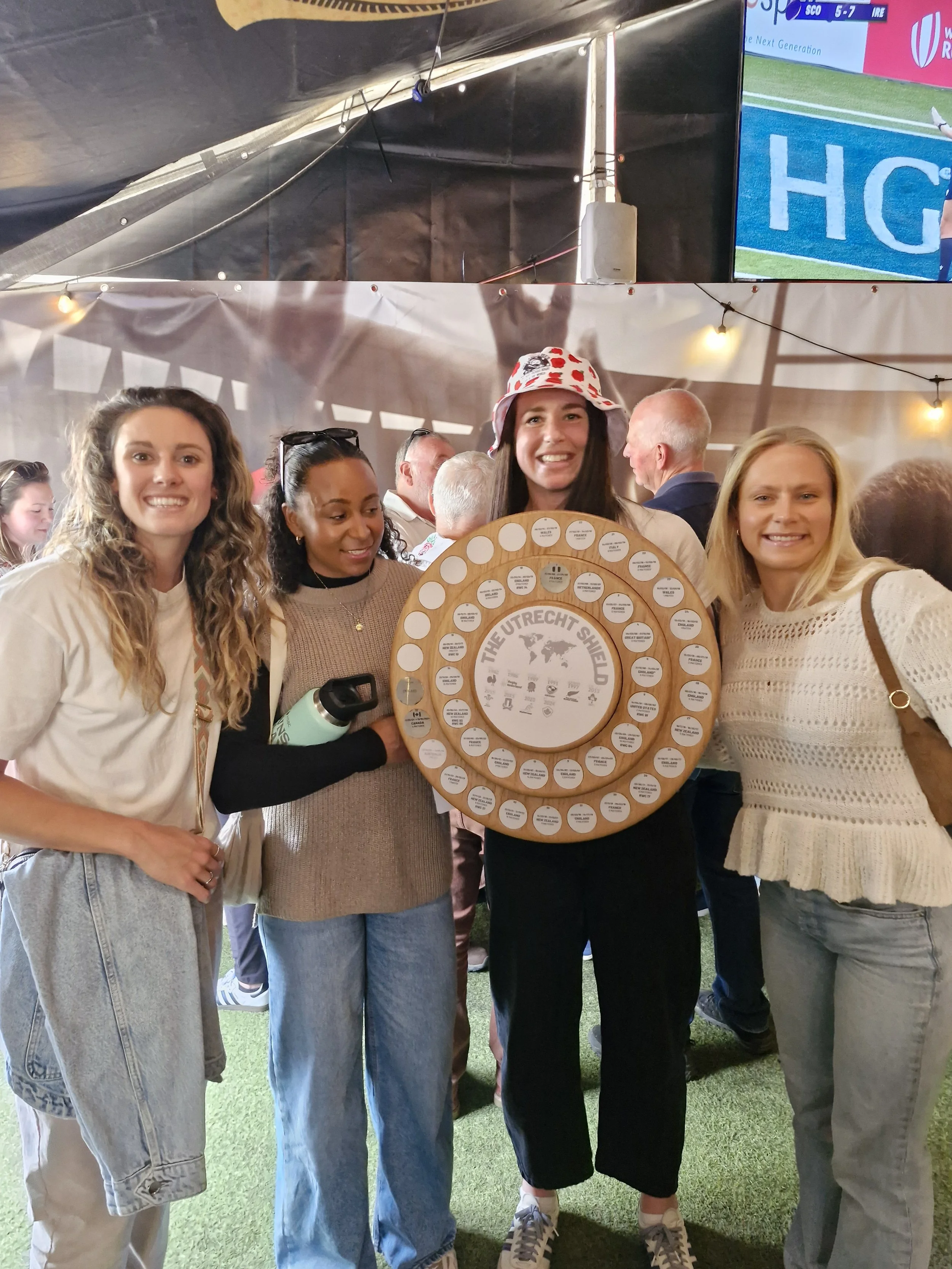 Group of four women smiling, standing indoors; one woman holds a circular award or plaque with the text 'The Utrecht Shield' and a world map at the center.