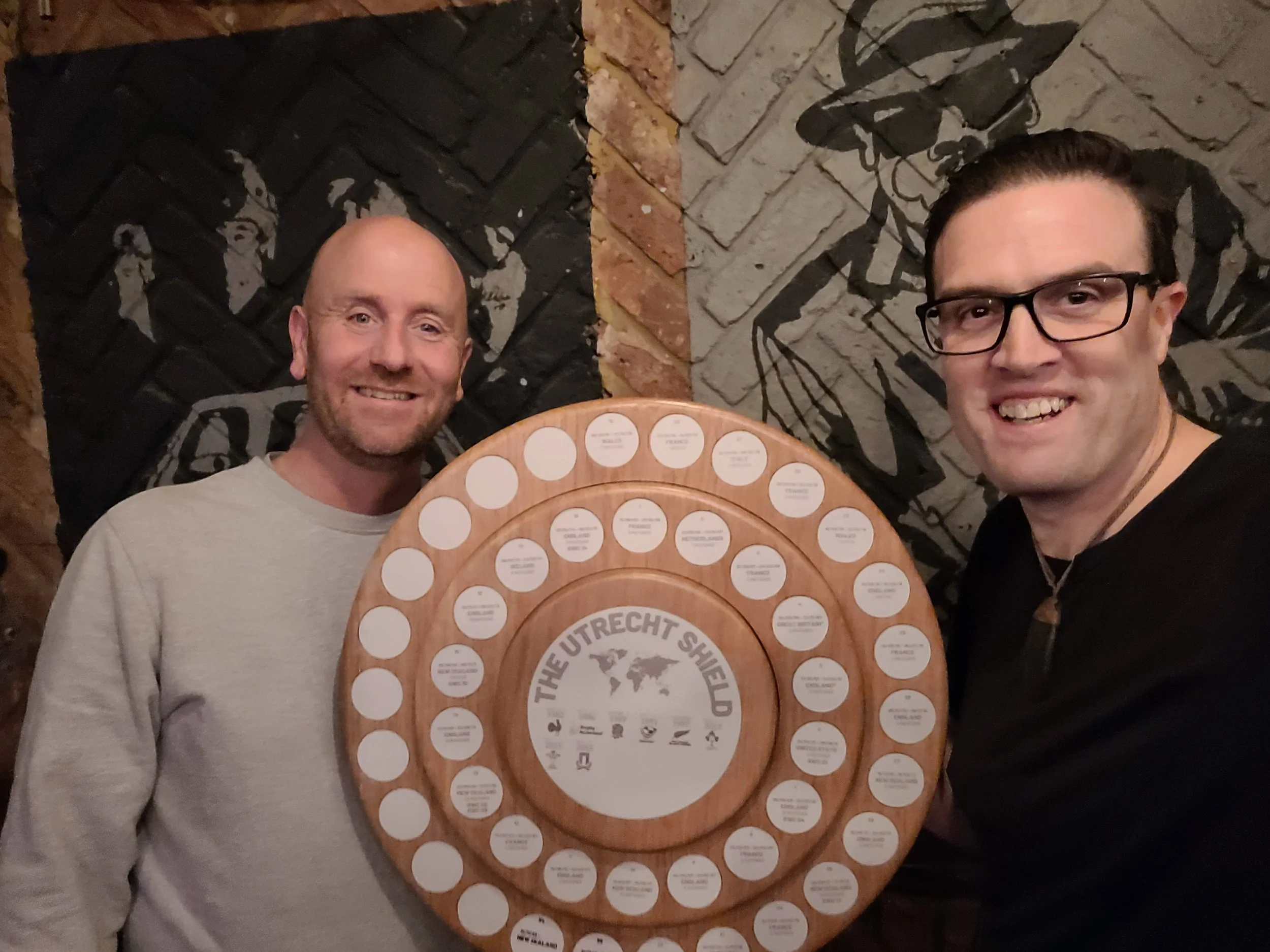 Two men smiling and posing for a photo with a circular wooden display board that has multiple small white circles with text. The background features a wall with graffiti-style artwork of two faces.