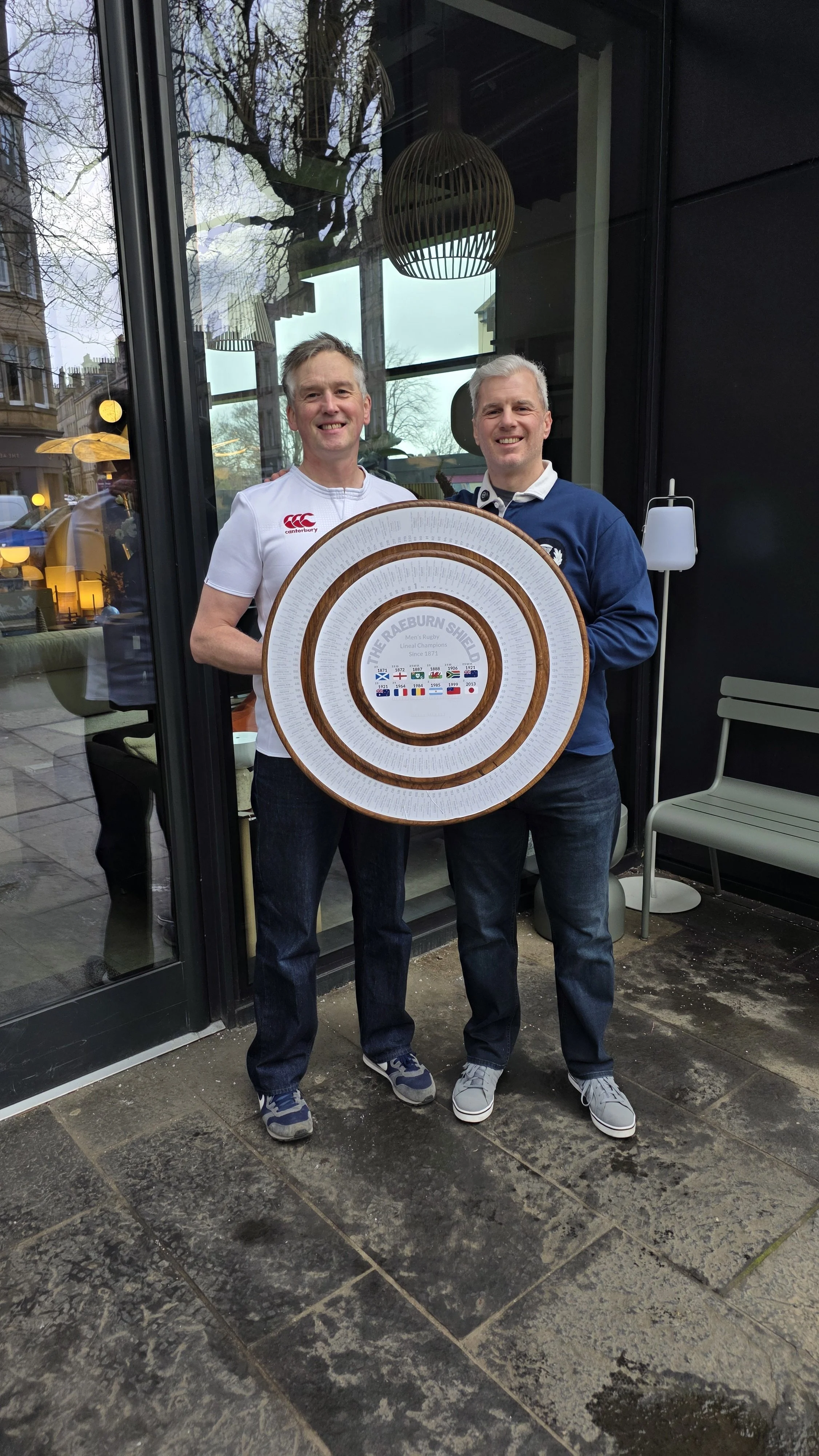 Two men standing outside a glass door, holding a large circular award or commemorative plaque with text and flags, smiling at the camera.