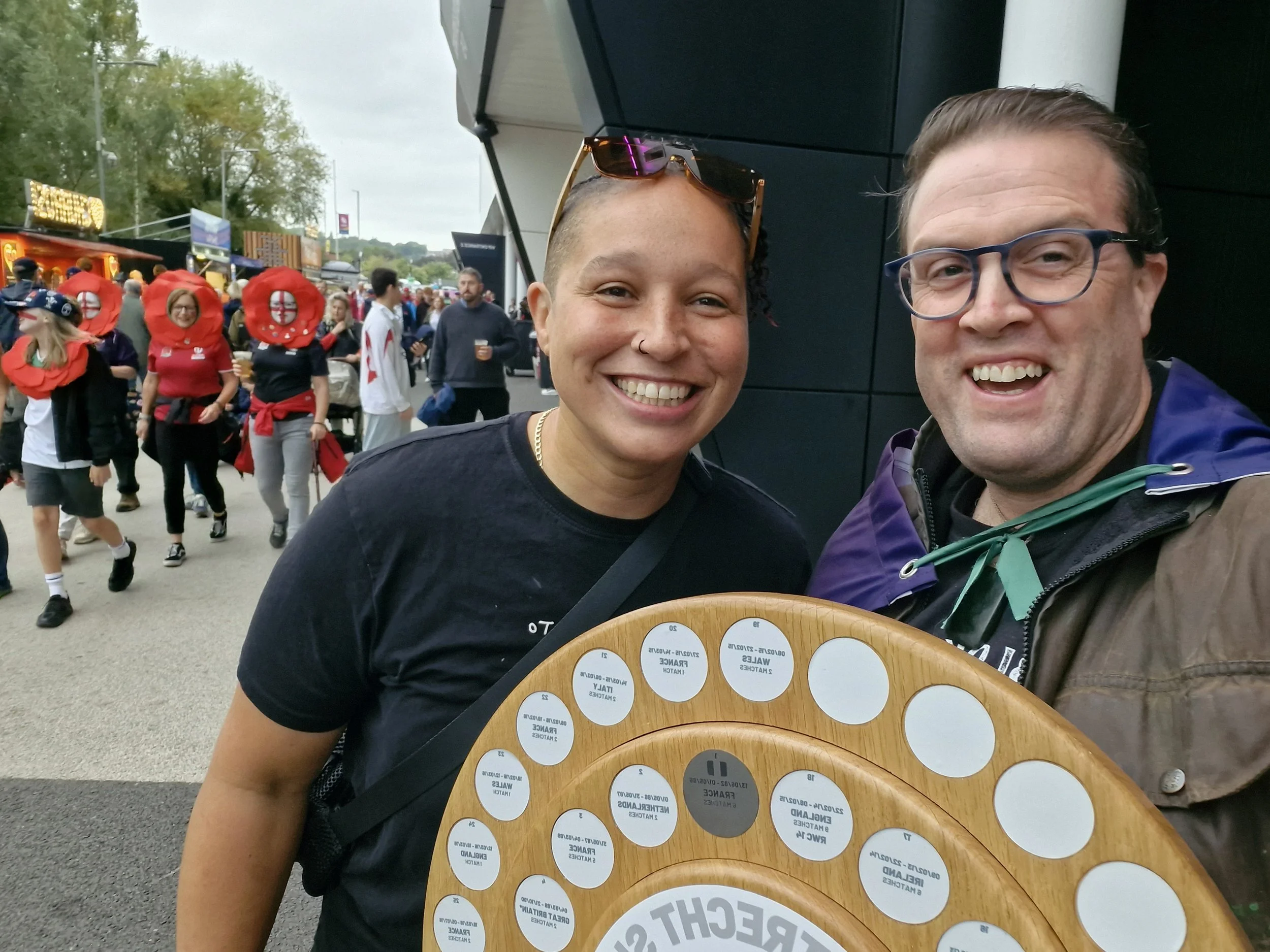 Two smiling people at a sports event holding an award board. In the background, people in mascot costumes and various attendees are visible at an outdoor venue.