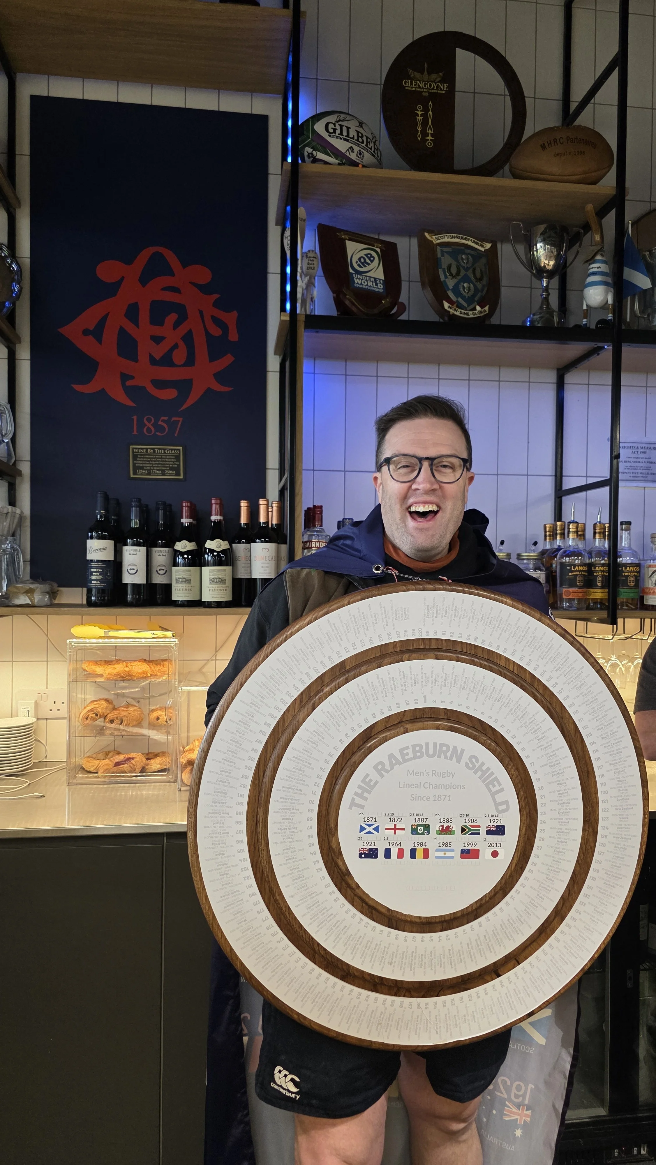 A man holding a large circular award plaque in a bar or pub setting, with shelves of wine bottles and trophies in the background.