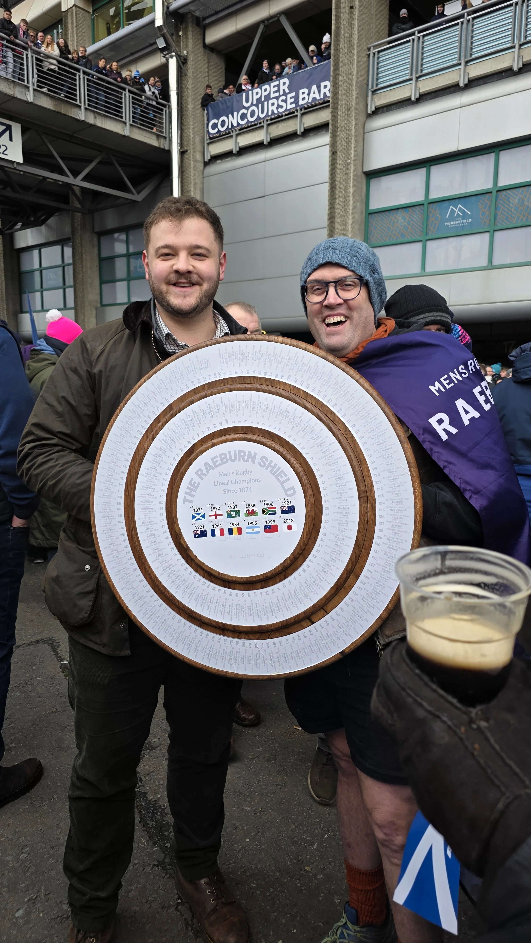 Two men smiling and holding a wooden shield that displays the history of The Raeburn Shield, with a crowd in the background at an outdoor event.
