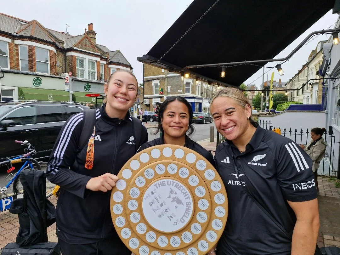 Three women in black uniforms holding a wooden wheel with white tags, standing on a city street with buildings and parked cars in the background.