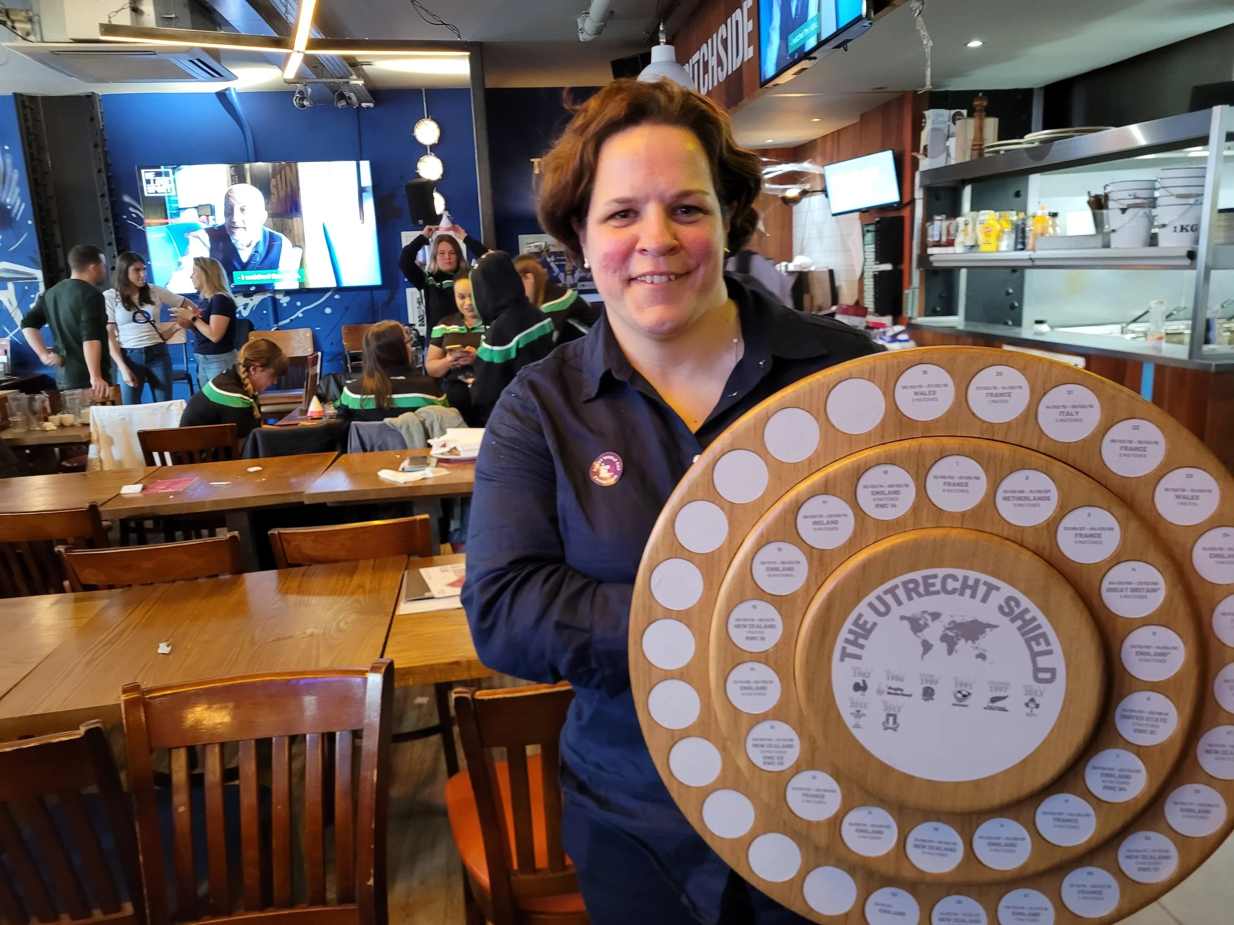 A woman holding a round wooden plaque with multiple small labels on it in a restaurant or sports bar setting. There are several people in the background, some sitting at tables and some standing, with a large television screen displaying a person and