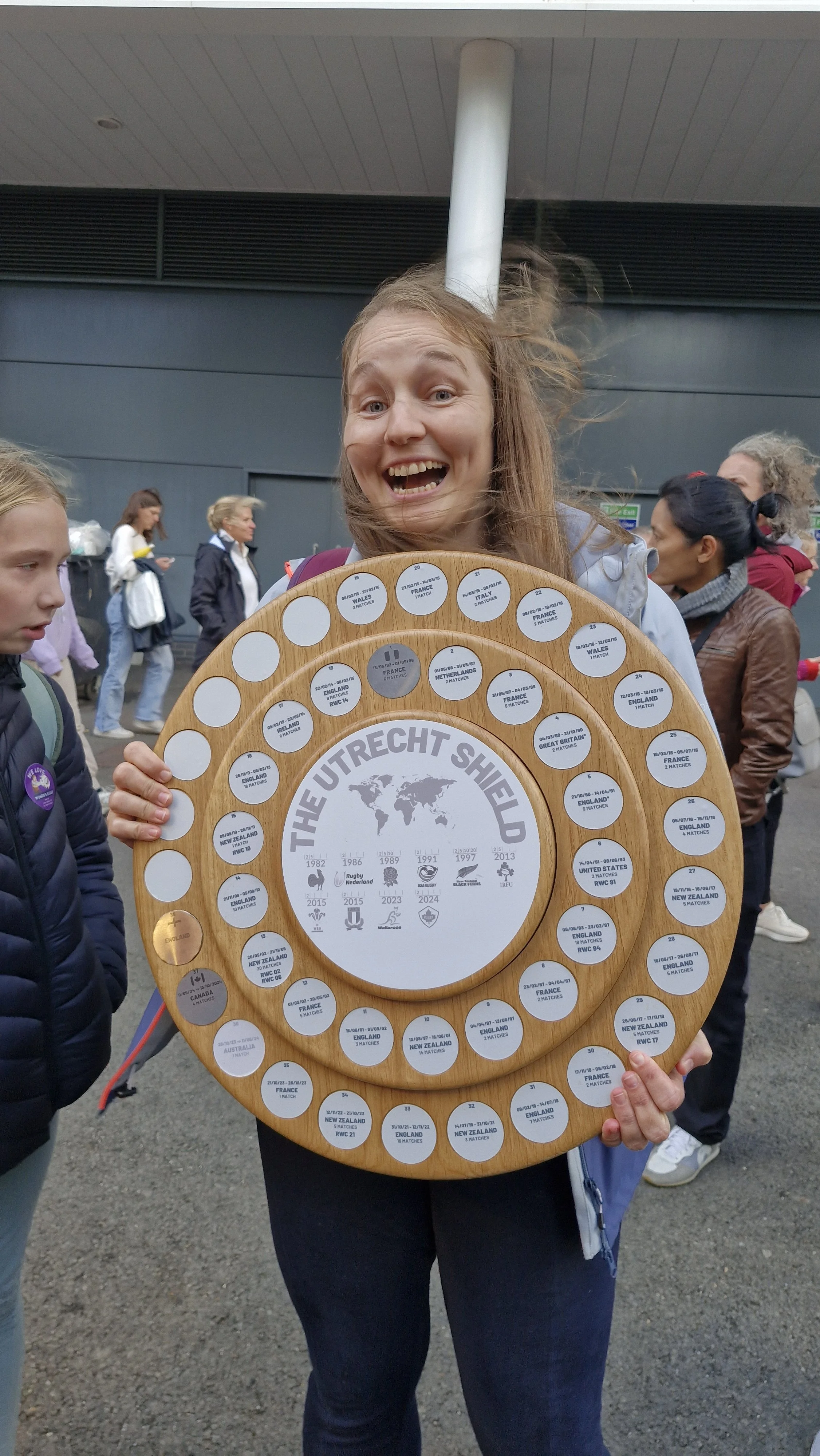 A woman happily holding a large wooden World Cup victory plaque, with several smaller plaques attached, showing matches between teams such as England, New Zealand, and France, at a stadium or outdoor event.