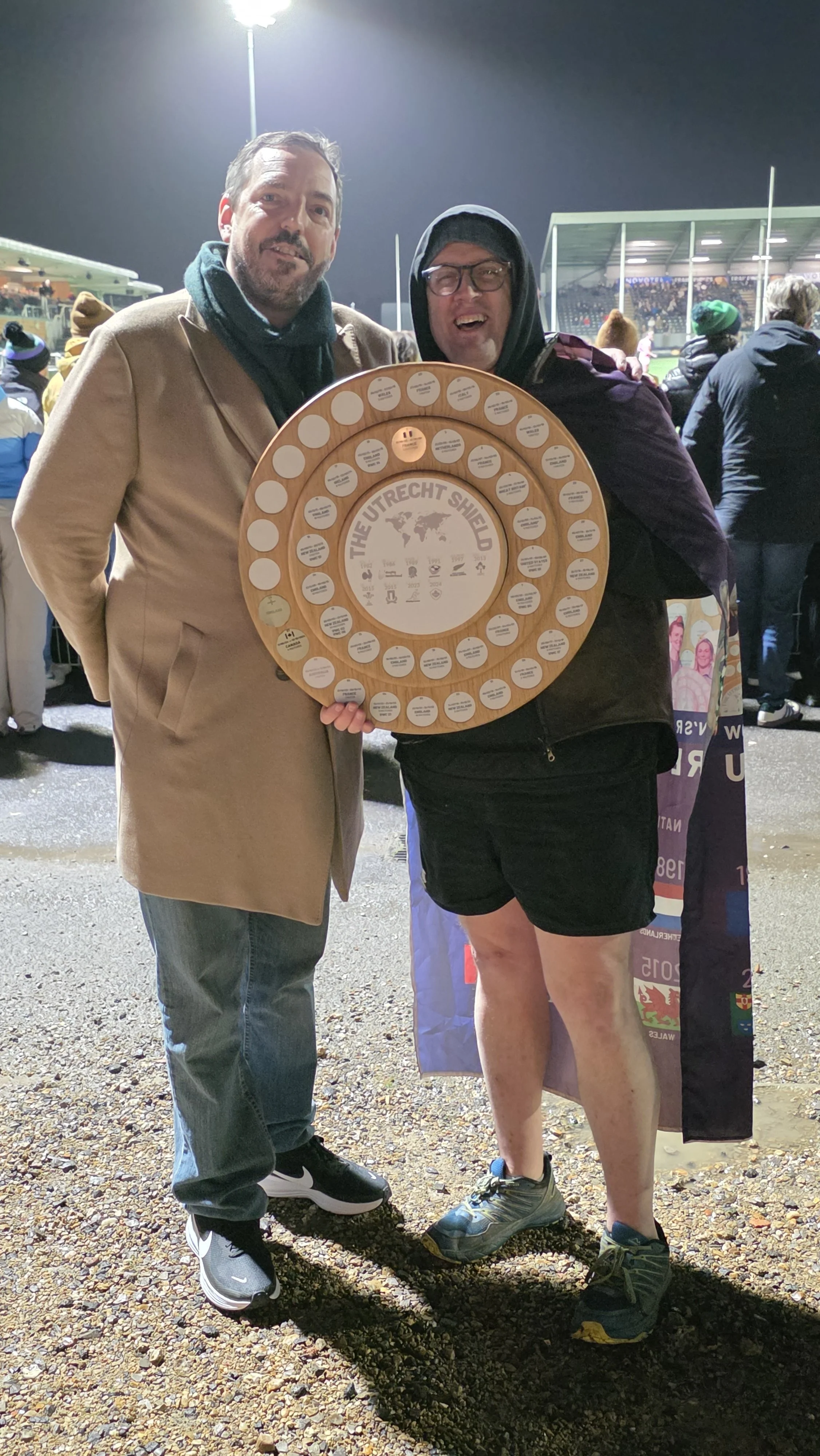 Two men standing outdoors at a sports event at night, one holding a circular wooden plaque with the words 'The Utrecht Shield' and various icons and text. The man on the left is wearing a beige coat and jeans, the man on the right is wearing a hoodie