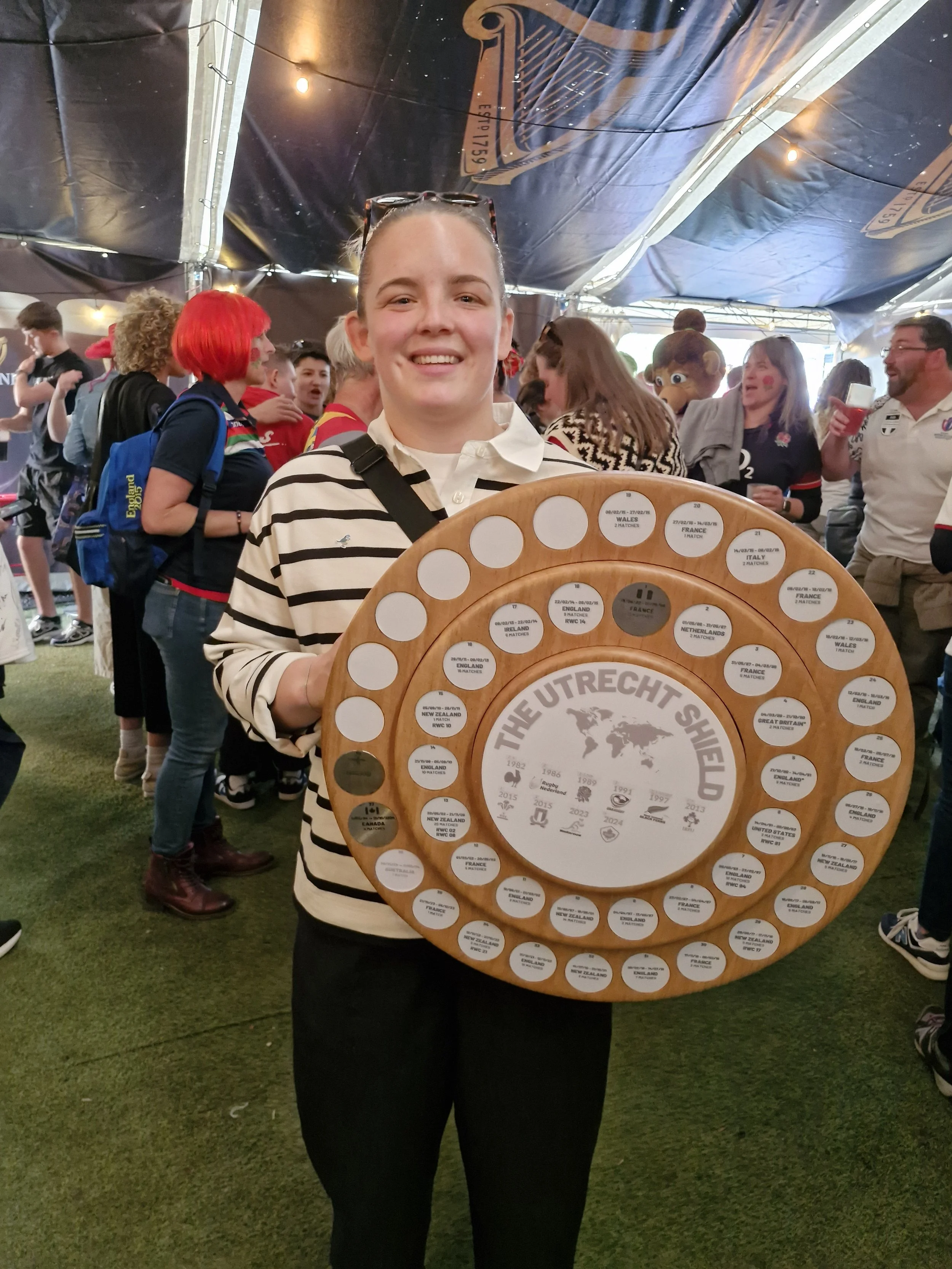 A smiling young woman holding a wooden plaque with multiple small circles, surrounded by a group of people in a tent, some wearing rugby jerseys and someone in a mascot costume.