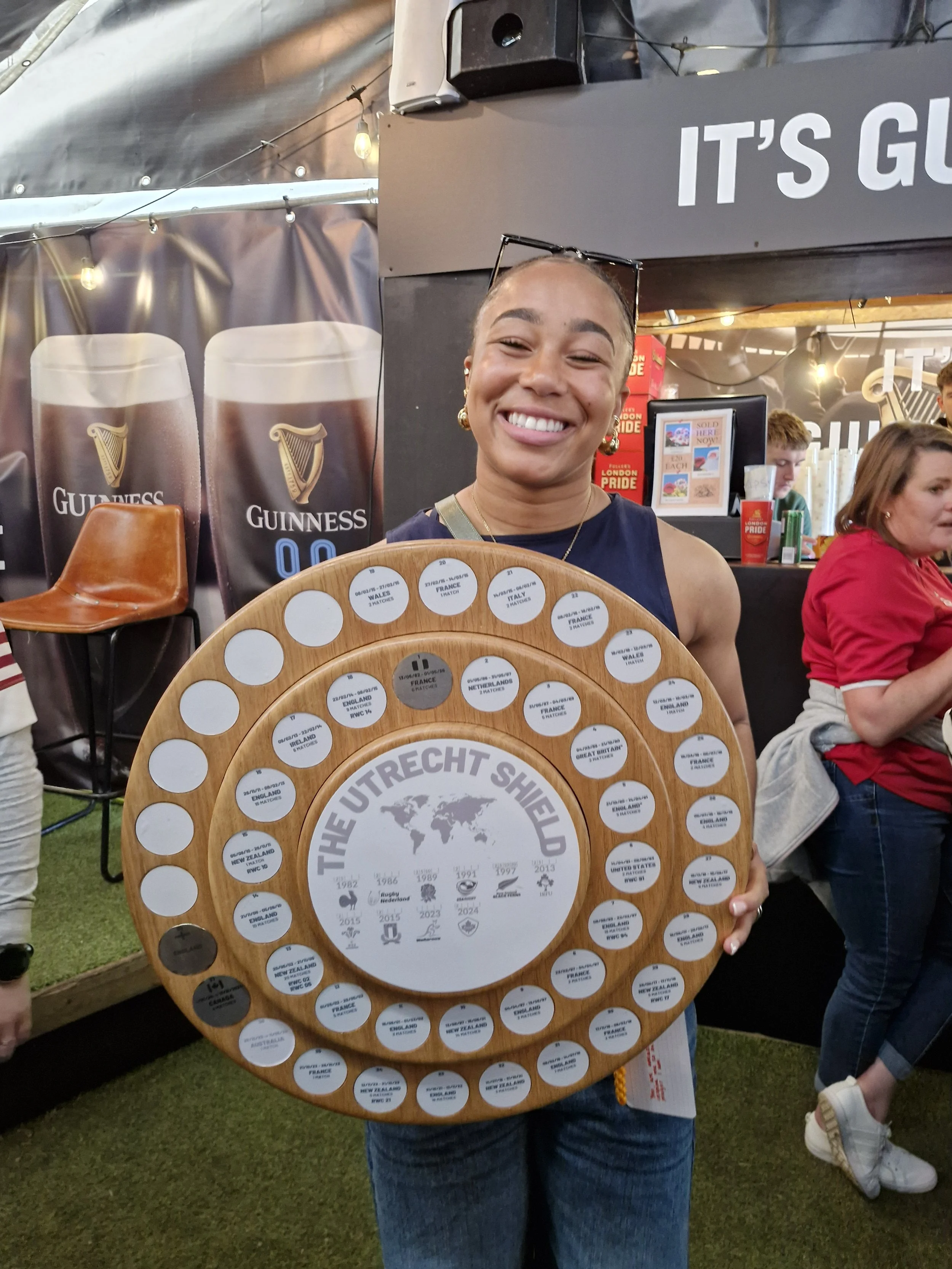 A woman smiling and holding a wooden board with flags of different countries and text reading 'The Utrecht Shield.' She is indoors at an event with Guinness posters and other people in the background.