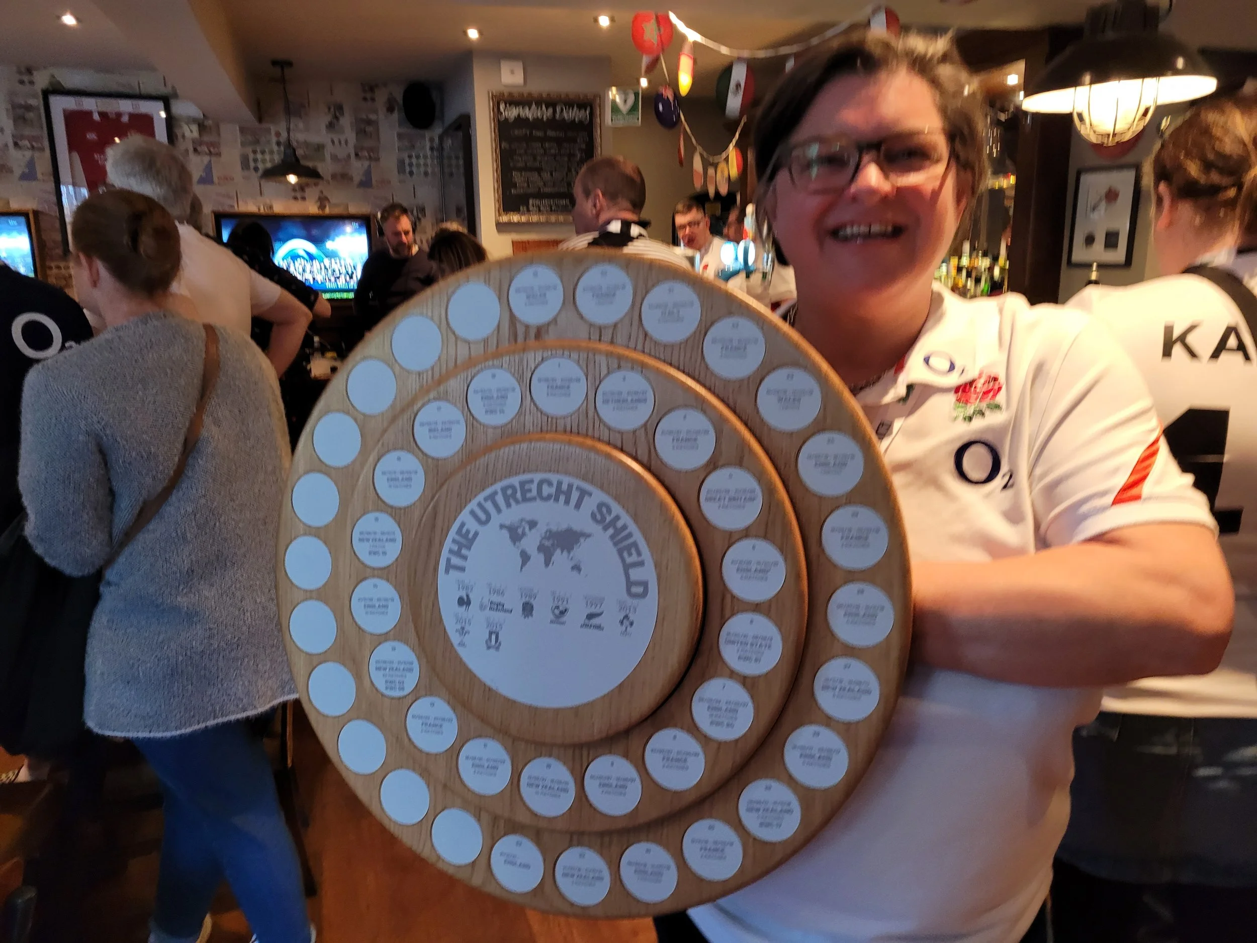 A woman wearing glasses and a white rugby shirt with the England rugby logo is smiling and holding a large circular wooden prize with small labels around its edge. The signs contain text related to The Utrecht Shield, and there are several people in 
