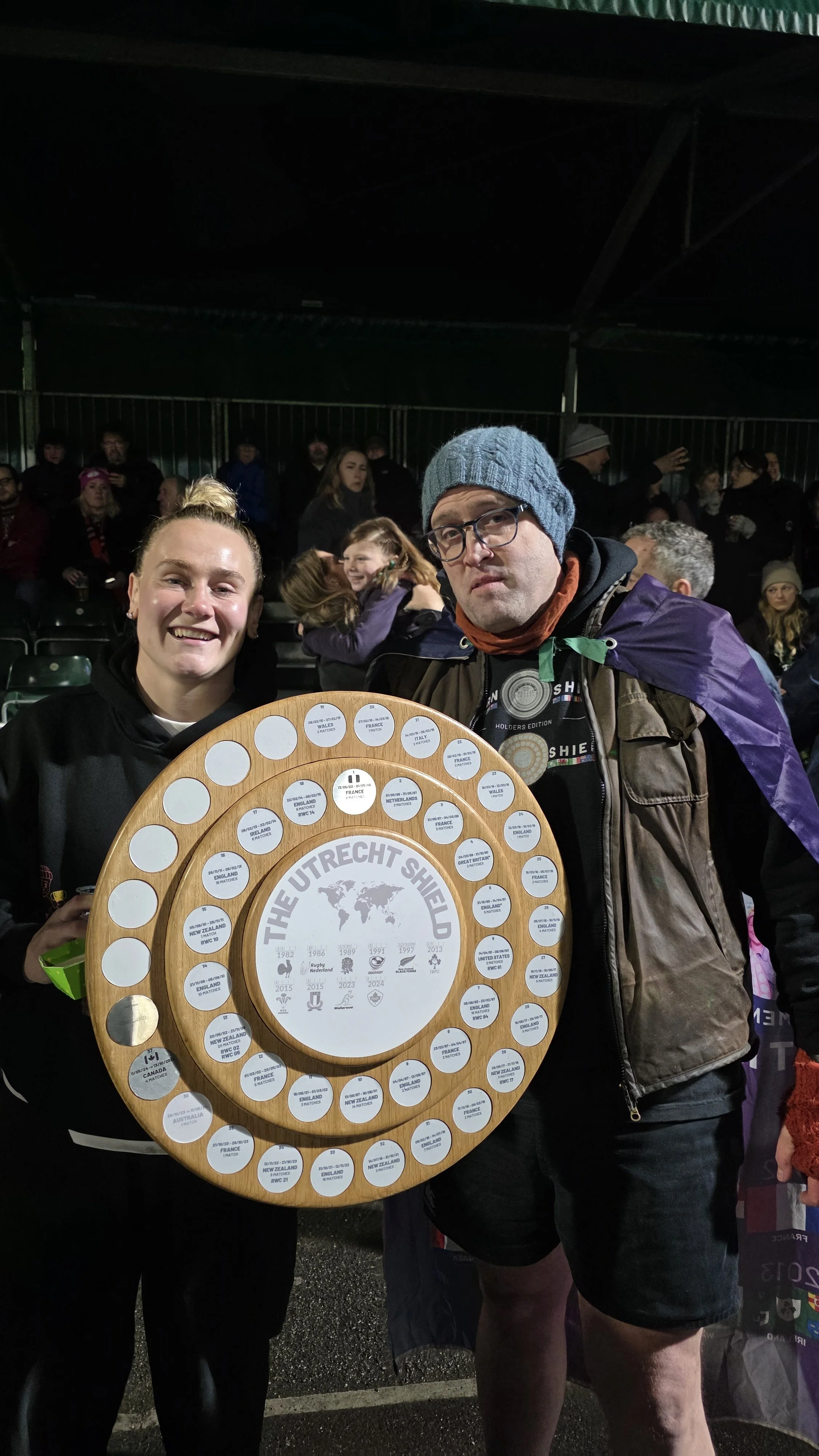 A smiling woman and a man wearing glasses and a beanie holding a wooden plaque with circular labels, at a night outdoor event, with other people in the background.