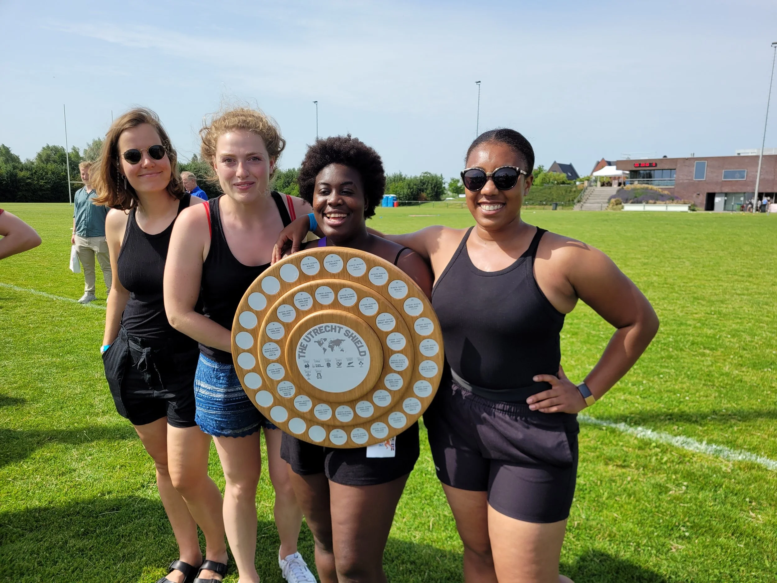 Four women standing on a grassy field holding a circular award plaque, smiling, with a building and other people in the background.