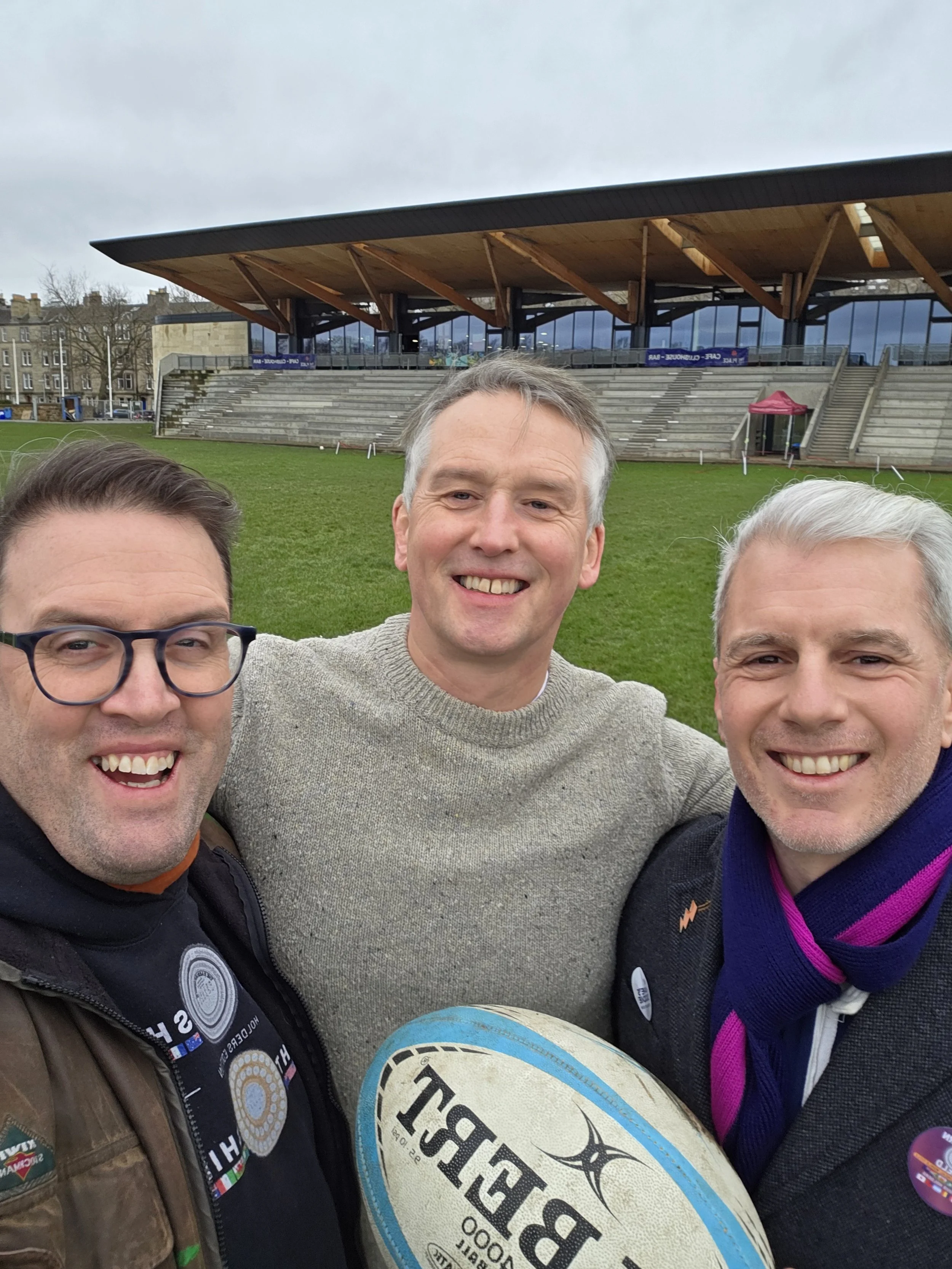 Three men smiling and taking a selfie on a sports field, with bleachers and a modern building in the background, and one holding a rugby ball.