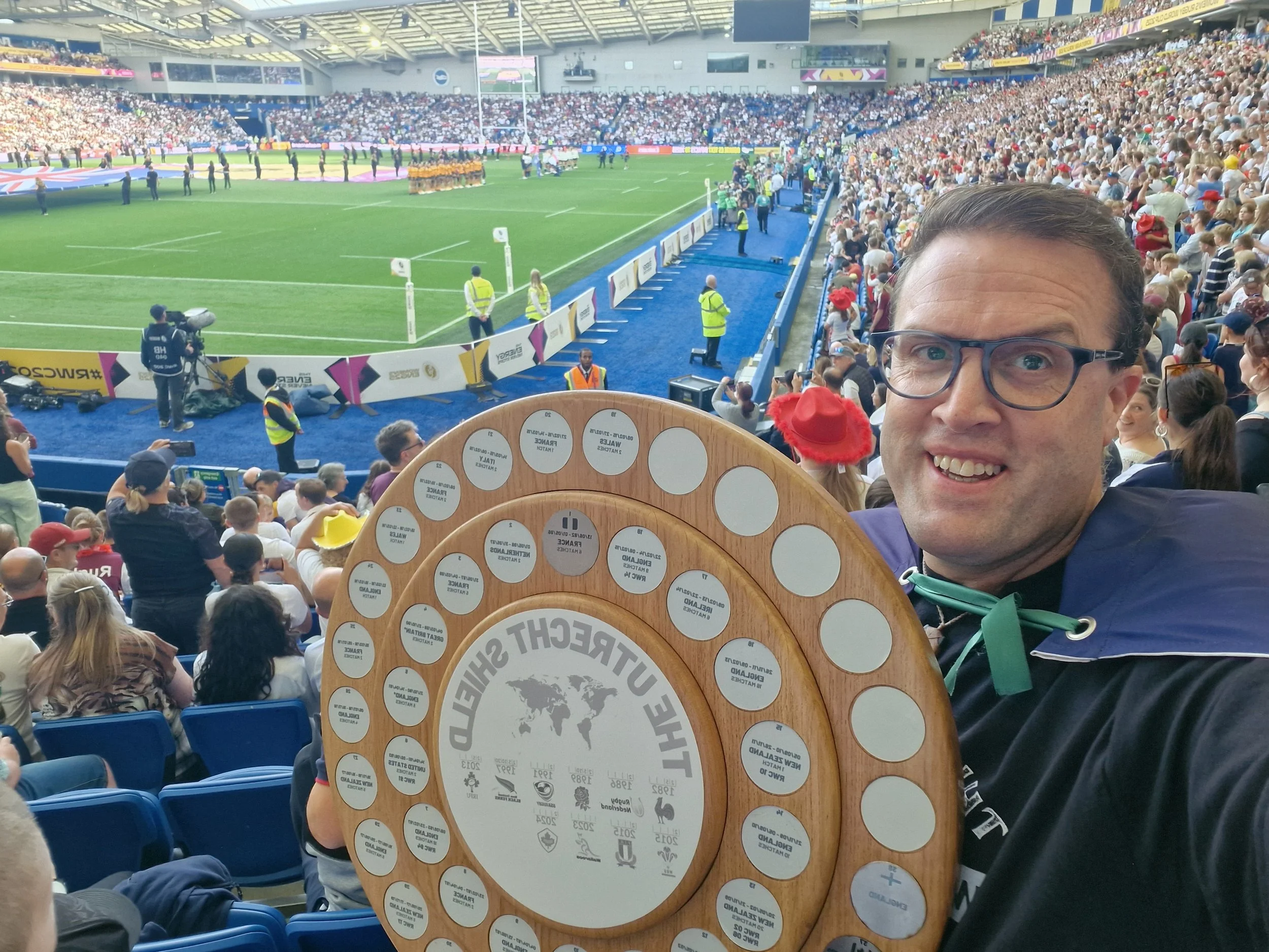 A man smiling at a sports stadium holding a wooden plaque with multiple small round tokens. The stadium is filled with spectators and a rugby match is underway on the field.