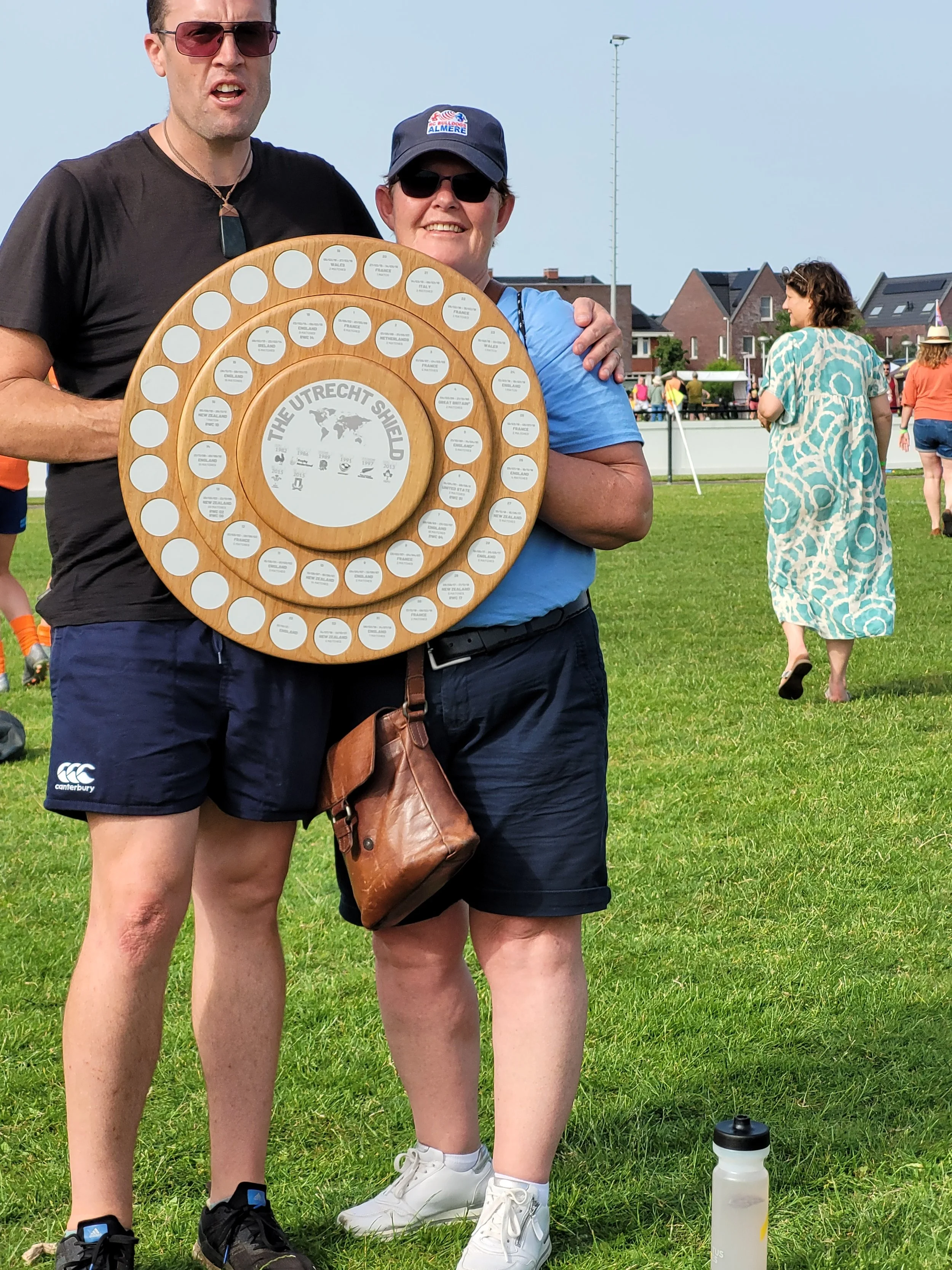 Two people, a man and a woman, standing on grass holding a wooden circular award with multiple labels. The man is wearing sunglasses, a dark T-shirt, and shorts, while the woman is wearing sunglasses, a cap, and a blue shirt. They are outdoors with s