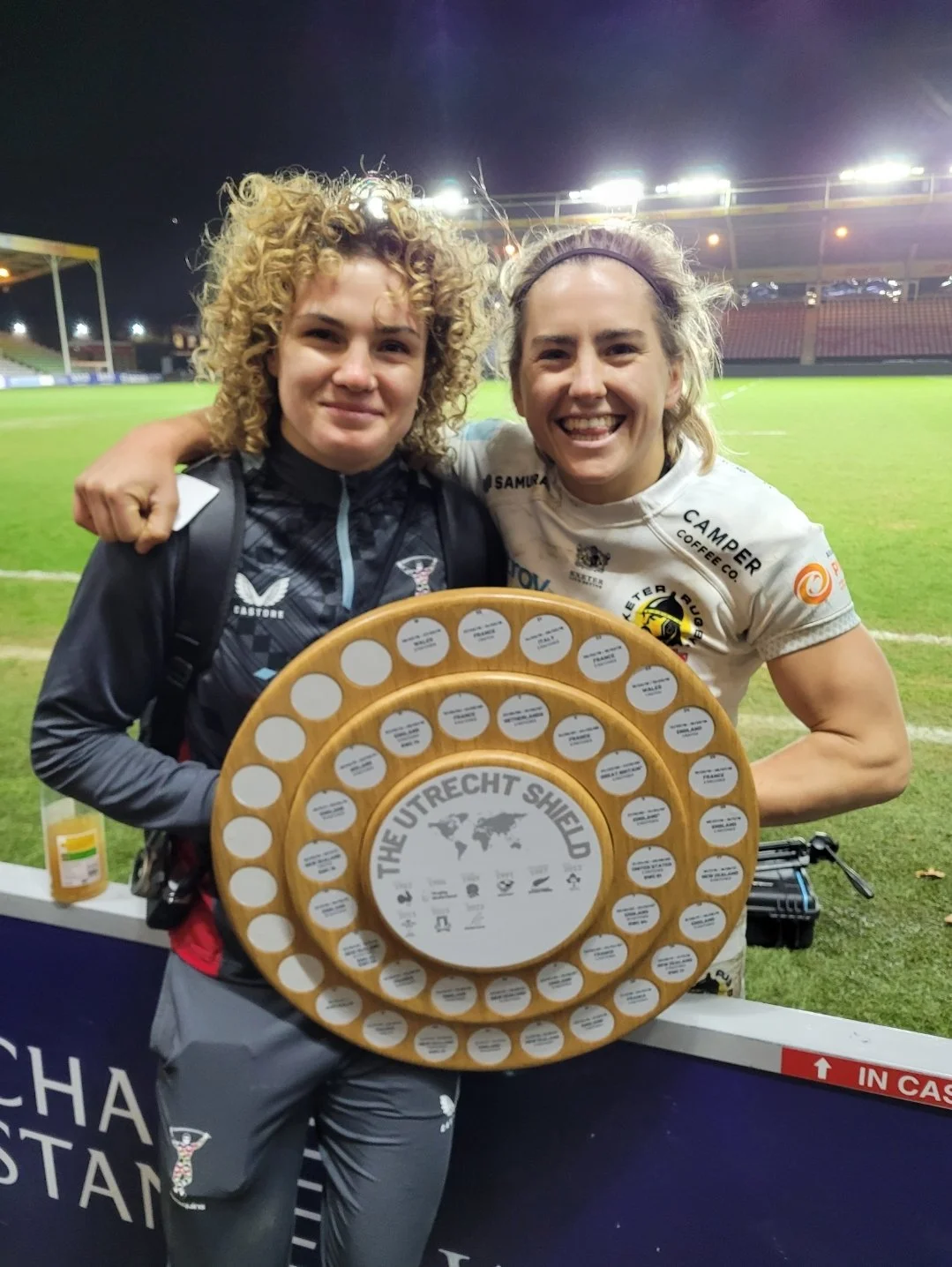 Two women celebrating on a sports field at night, holding a large circular award with multiple smaller labels, smiling with arms around each other.