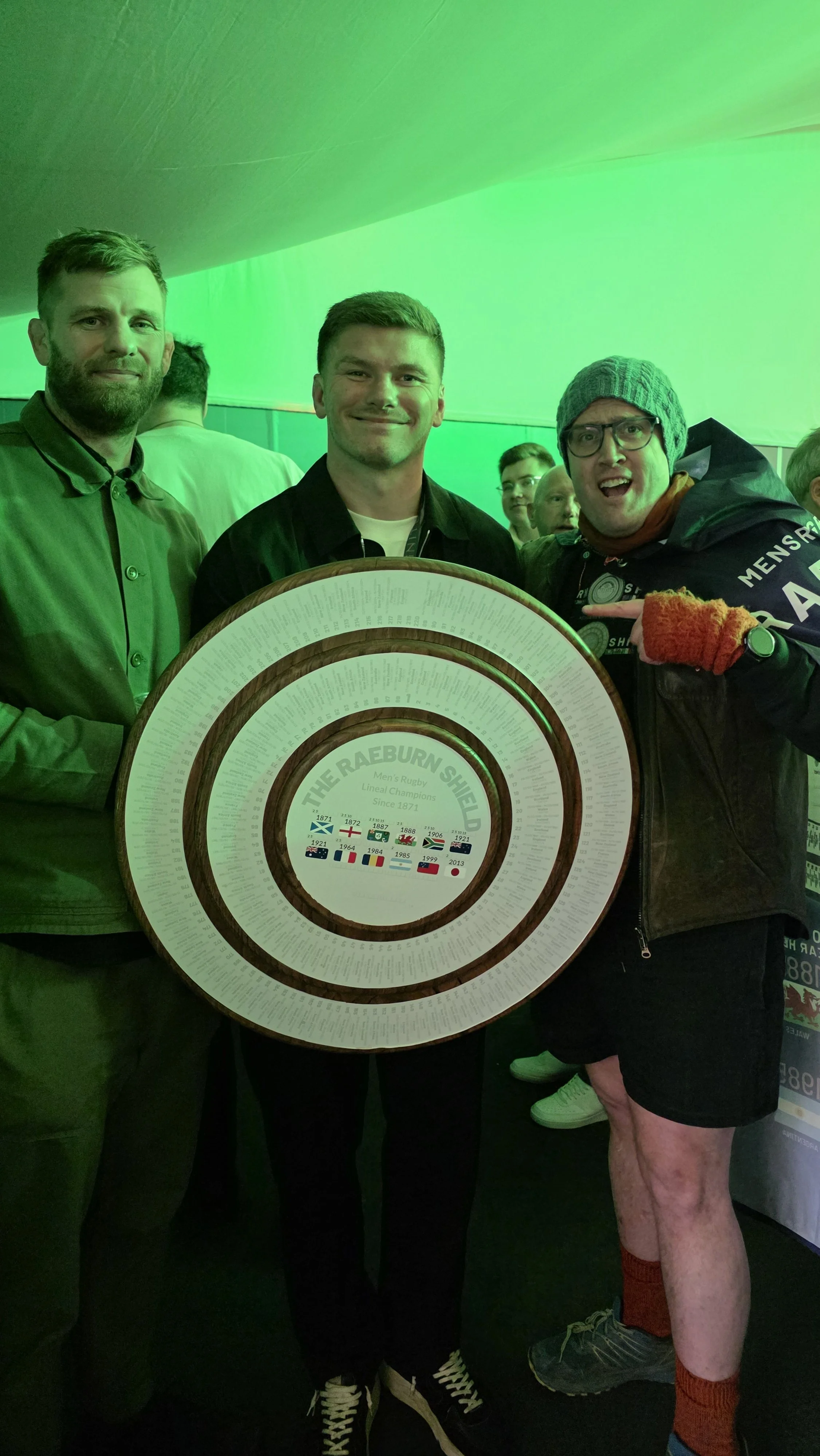 Three men standing indoors under green lighting, one holding a large circular commemorative plaque for The Raeburn Shield with Scottish flag and rugby history, others smiling and pointing, with other people visible in the background.