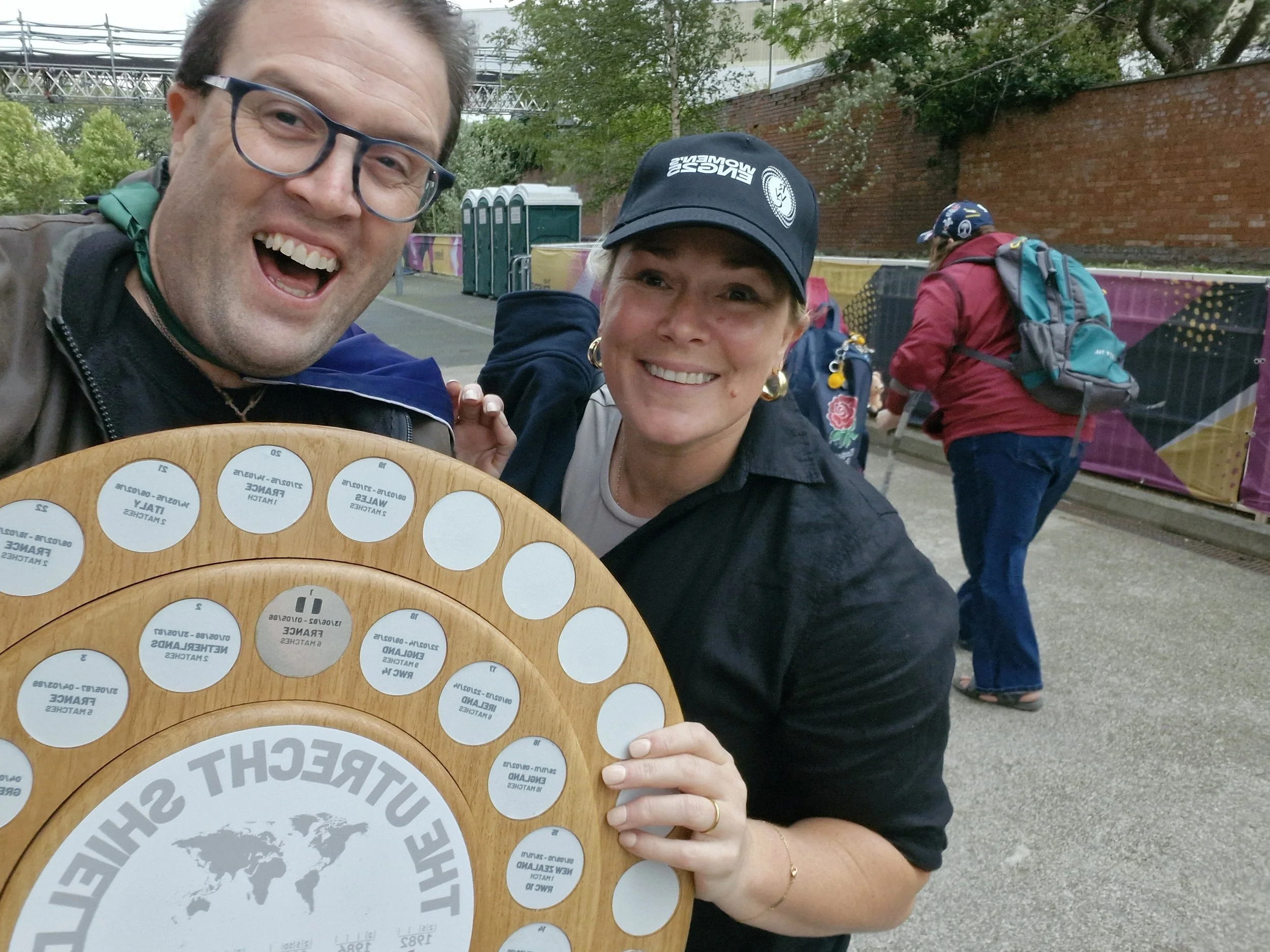 Two smiling people holding a UNICEF World Children's Day award plaque outdoors, with other people and portable toilets in the background.